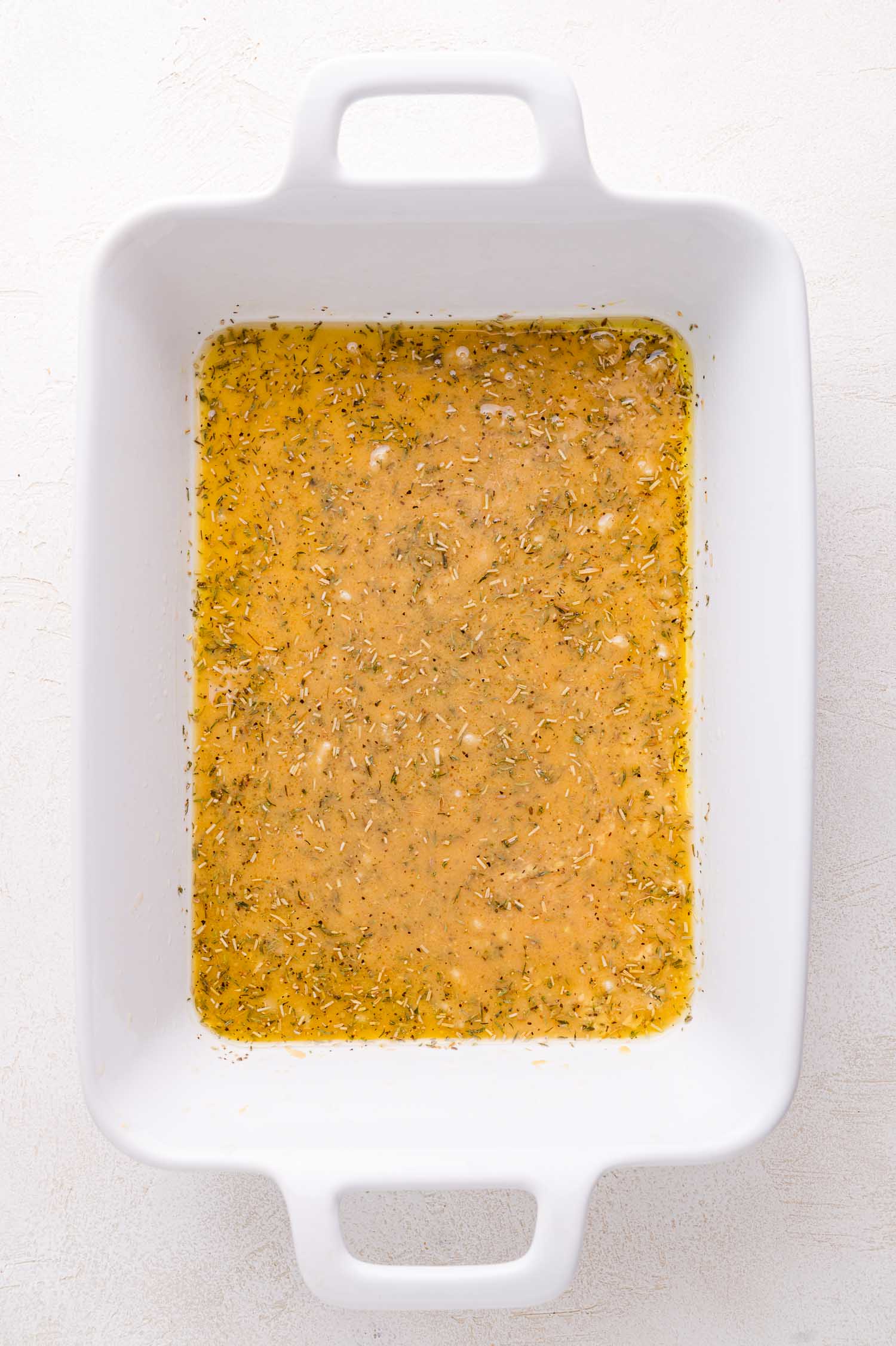 White rectangular baking dish filled with a yellowish marinade containing herbs and spices, placed on a light-colored surface.