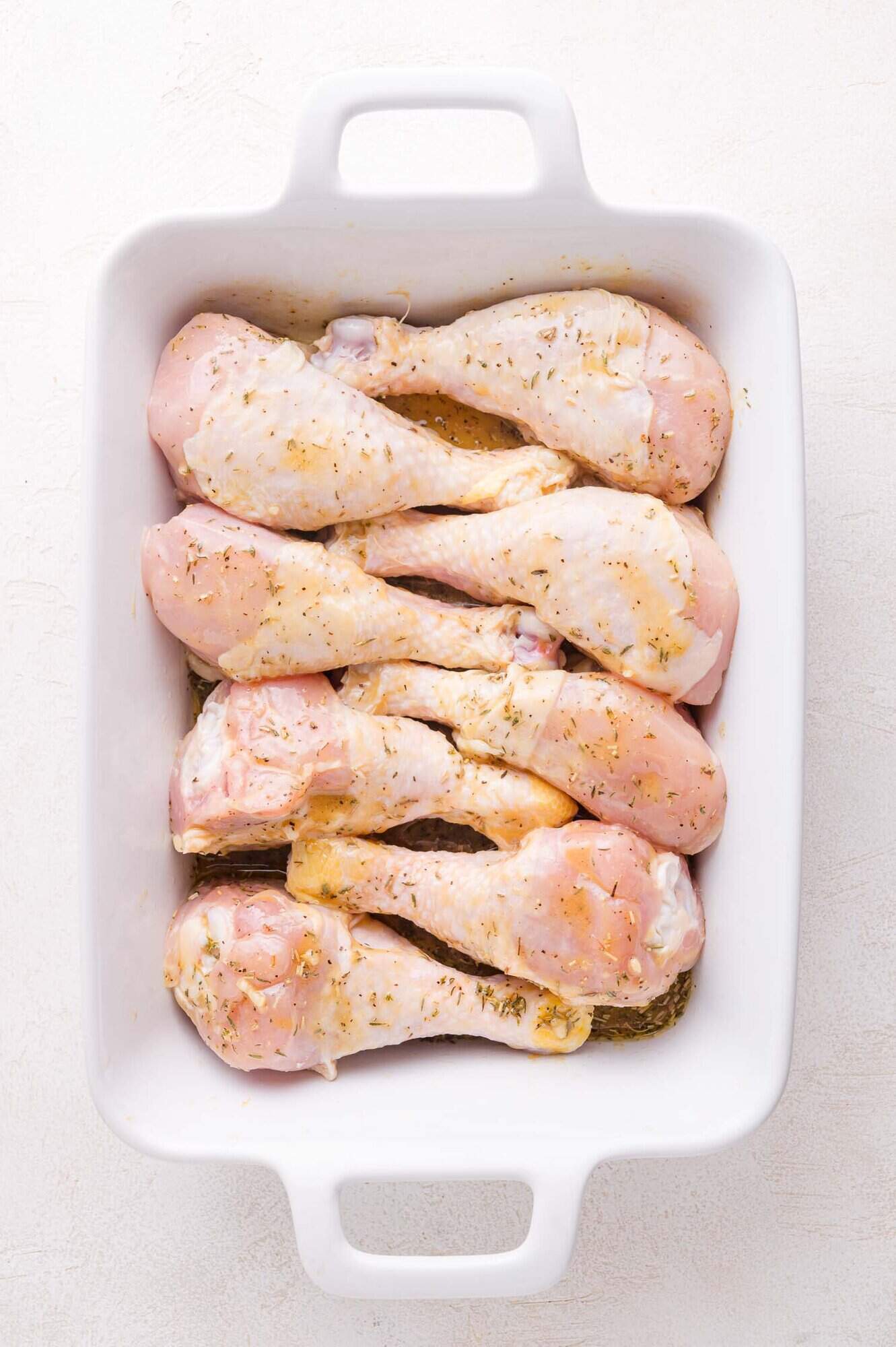 Raw, seasoned chicken drumsticks marinating in a single layer inside a white rectangular baking dish on a light surface.