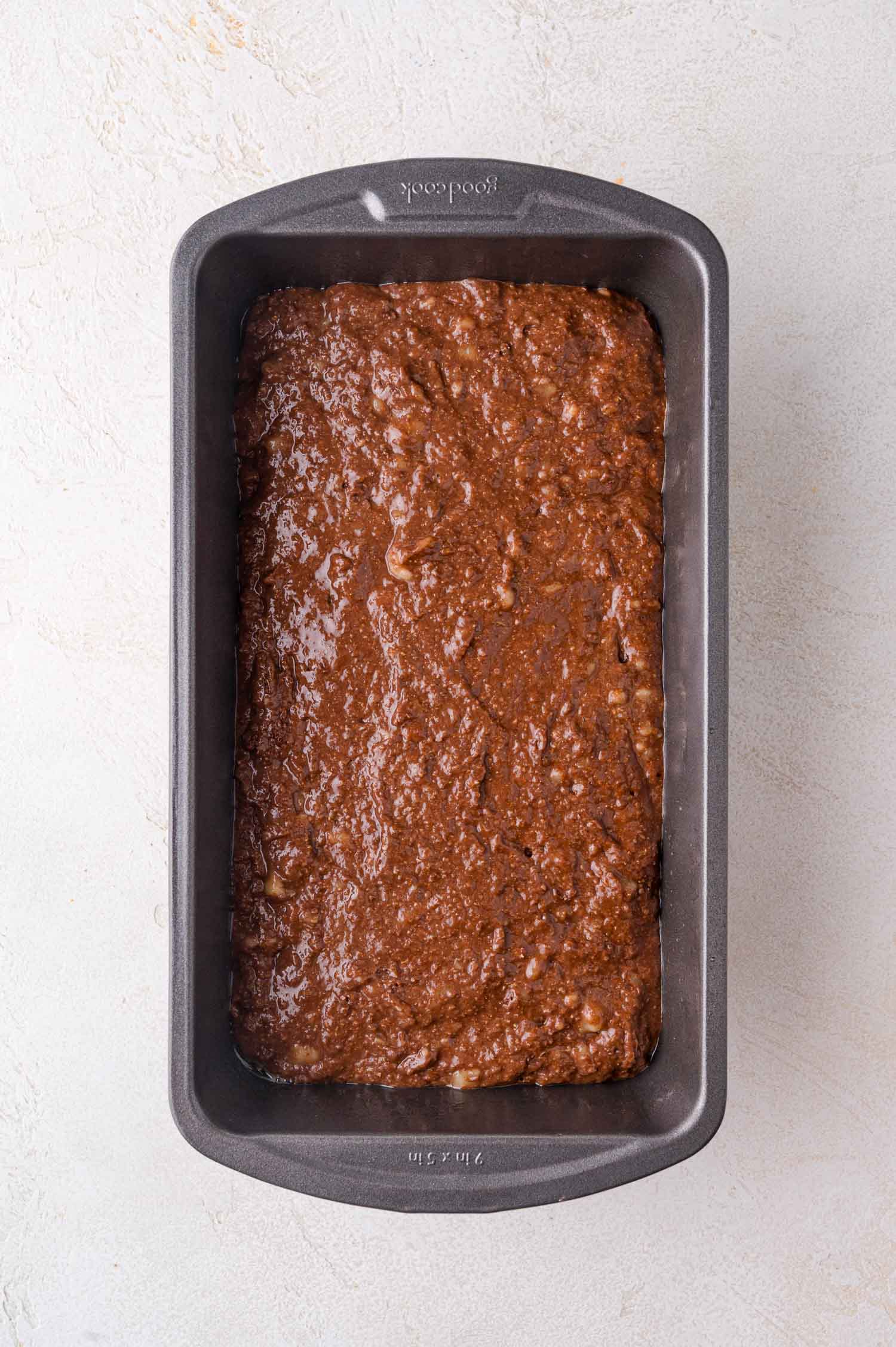 A loaf pan filled with unbaked chocolate banana bread batter on a light-colored surface.