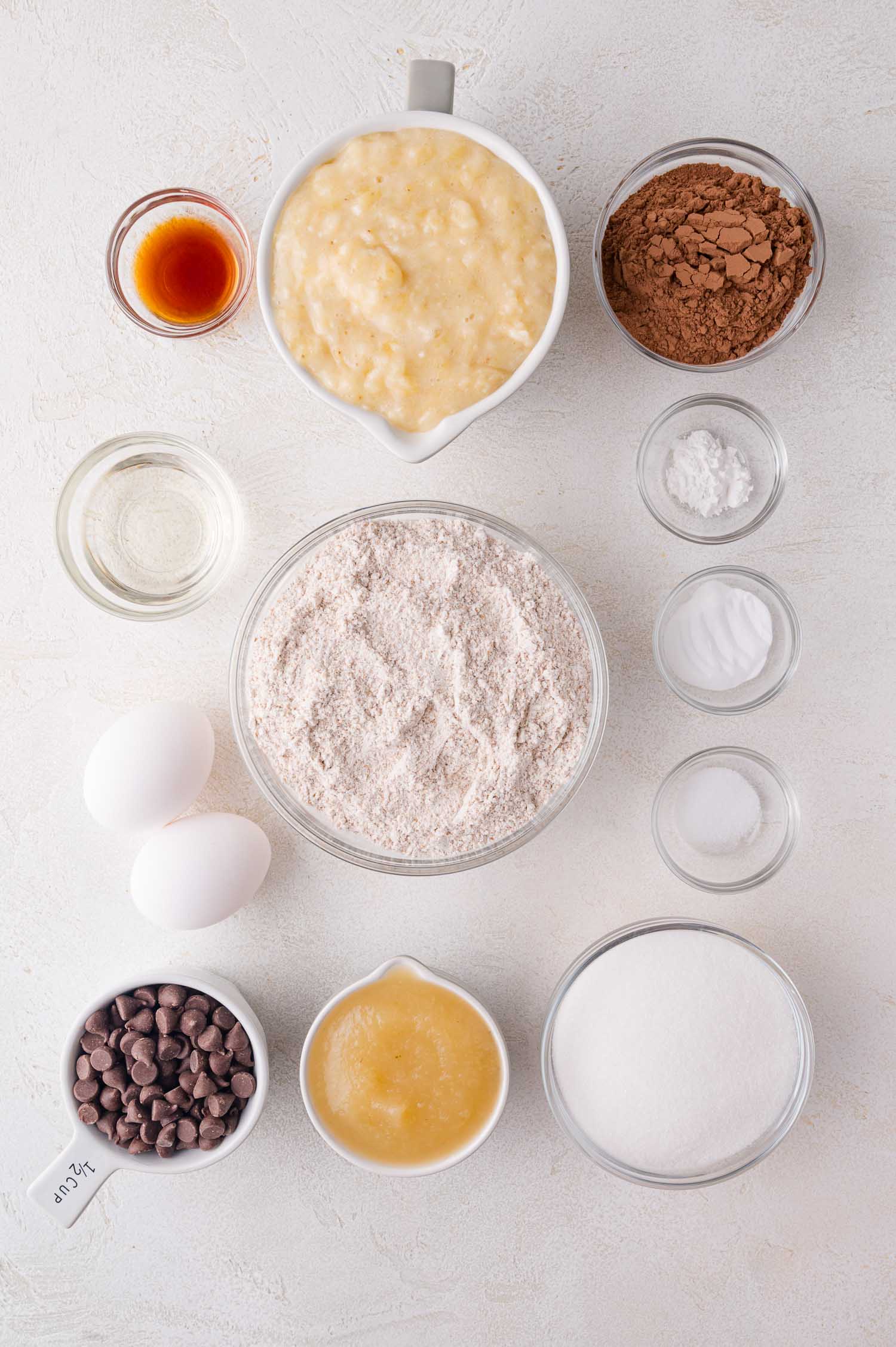 Top-down view of measured baking ingredients in bowls, including flour, mashed bananas, cocoa powder, eggs, sugar, chocolate chips, oil, applesauce, vanilla, and leavening agents.