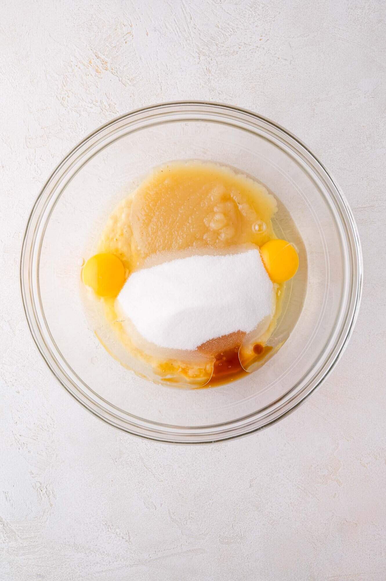 A glass bowl containing applesauce, two raw eggs, sugar, and vanilla extract on a light surface.