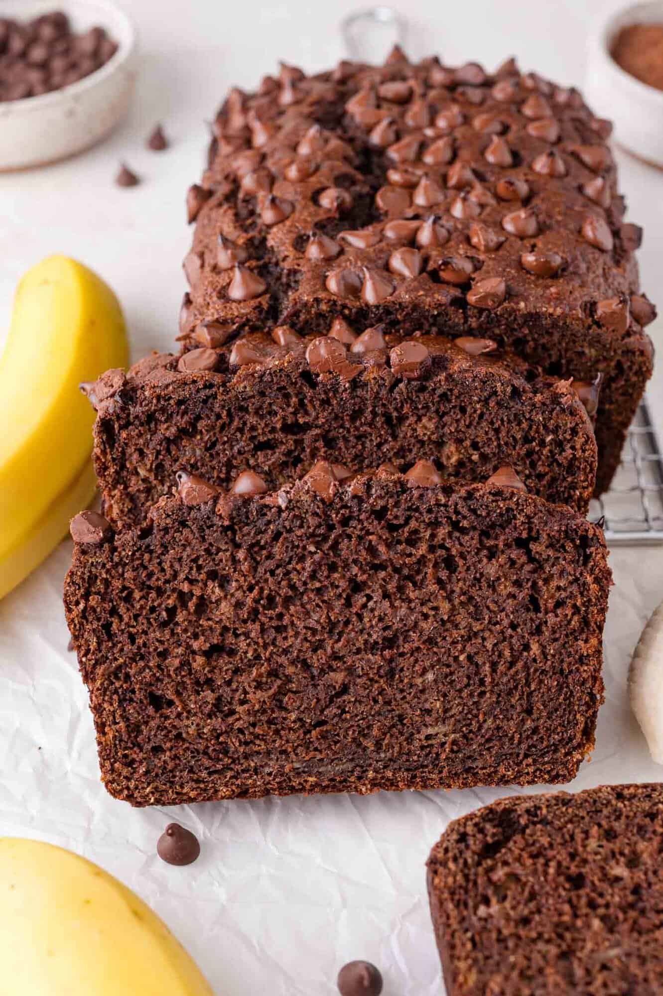 A sliced loaf of chocolate banana bread topped with chocolate chips, surrounded by bananas and chocolate chips on a light surface.