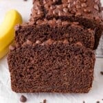 A close-up of sliced healthy chocolate banana bread with chocolate chips on top, next to a banana on a white surface.
