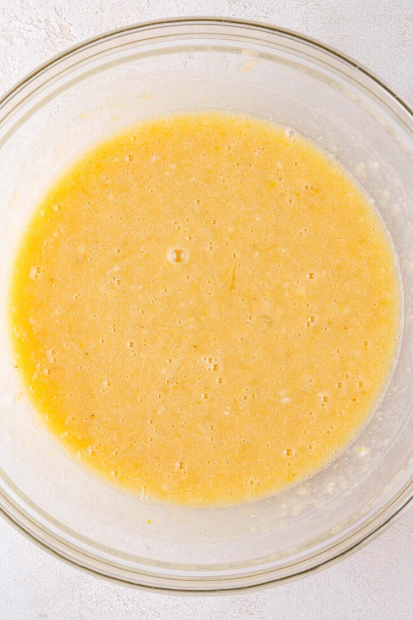 A glass bowl filled with a yellow, slightly lumpy batter mixture on a light-colored surface.