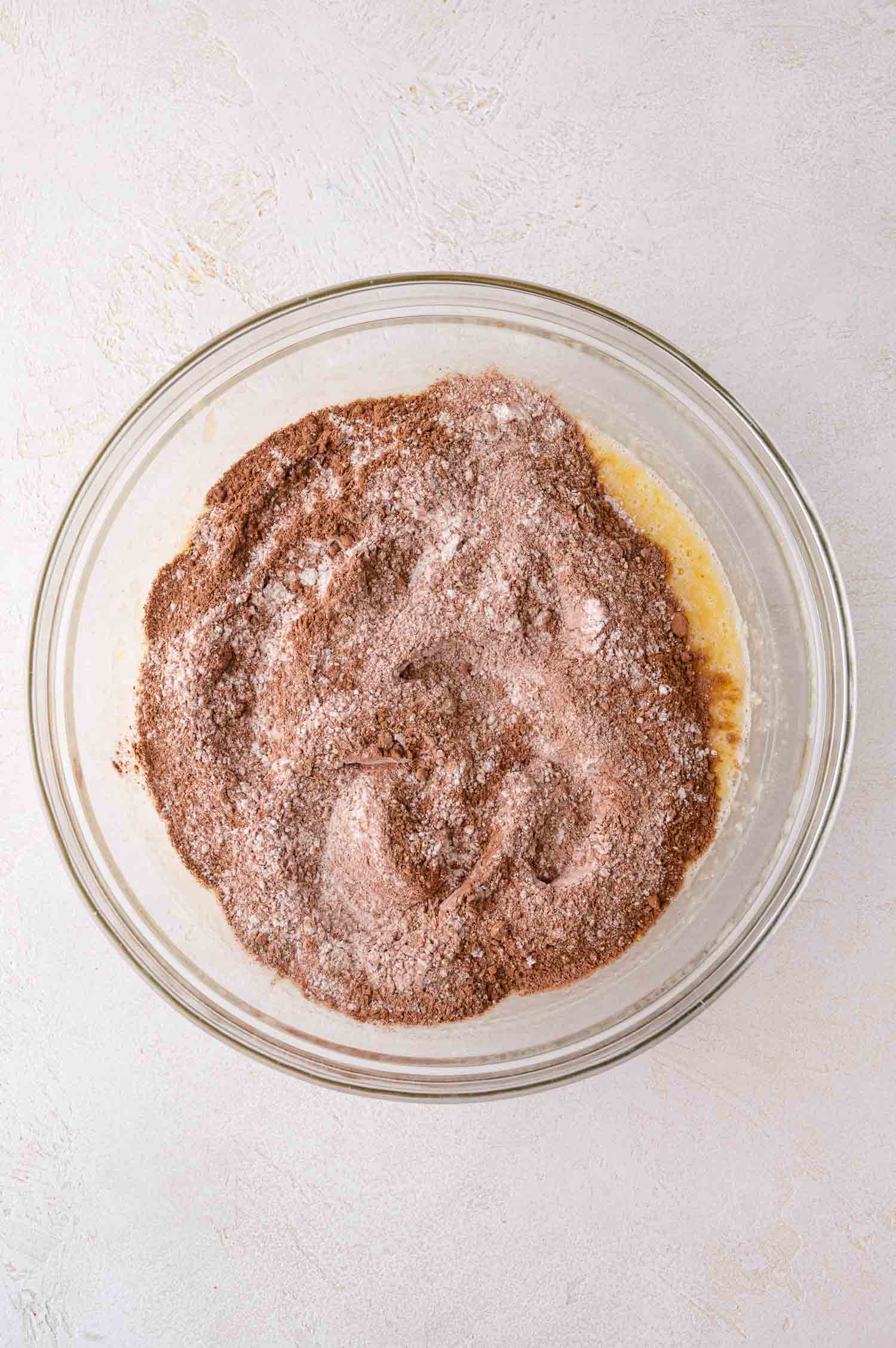 A glass bowl containing unmixed dry cocoa powder and flour on top of wet ingredients, viewed from above on a light surface.