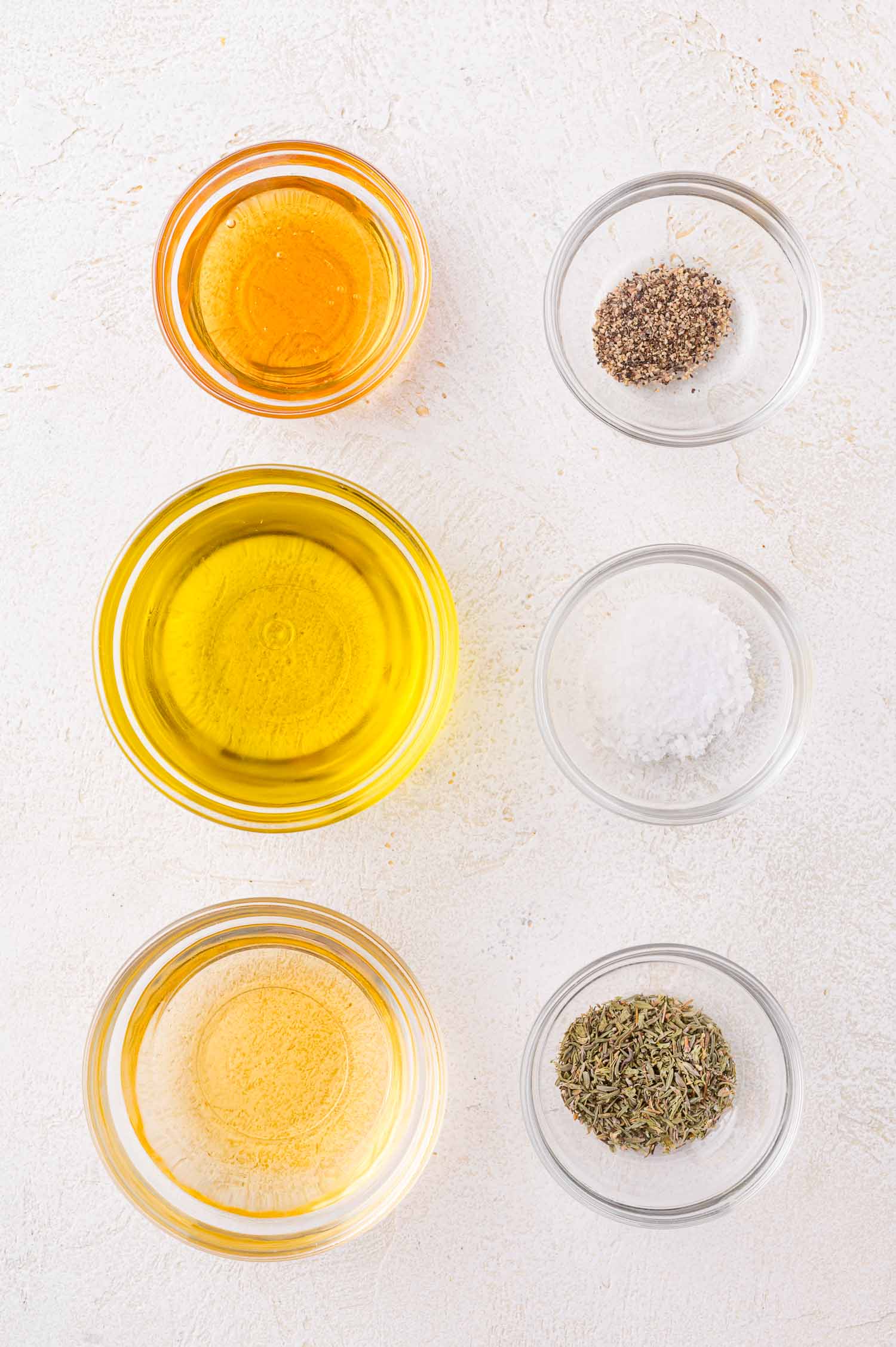 Six small glass bowls containing olive oil, vinegar, honey, dried herbs, salt, and pepper are arranged on a white surface.