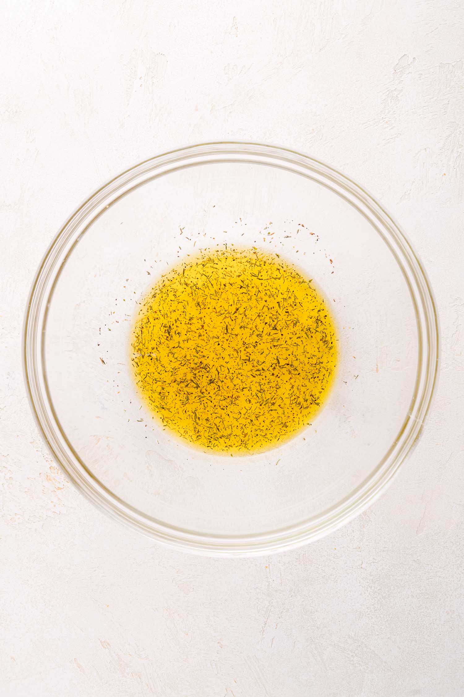 A glass bowl containing yellow salad dressing liquid with herbs mixed in, placed on a light-colored surface.