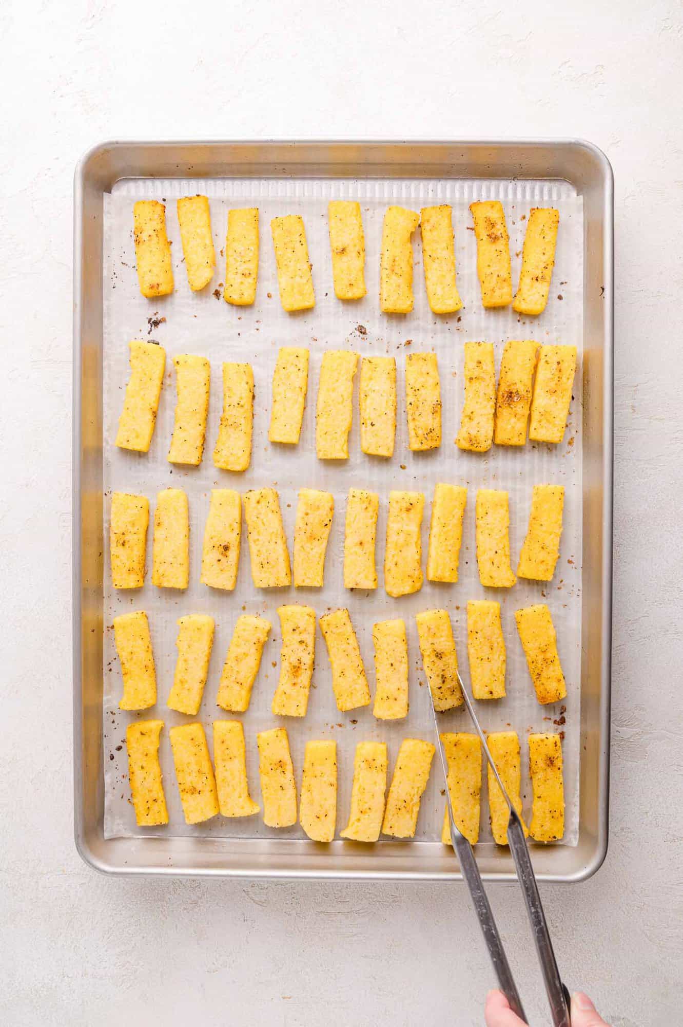 A baking tray lined with parchment paper holds evenly spaced, seasoned polenta fries, with a hand using tongs to arrange them.
