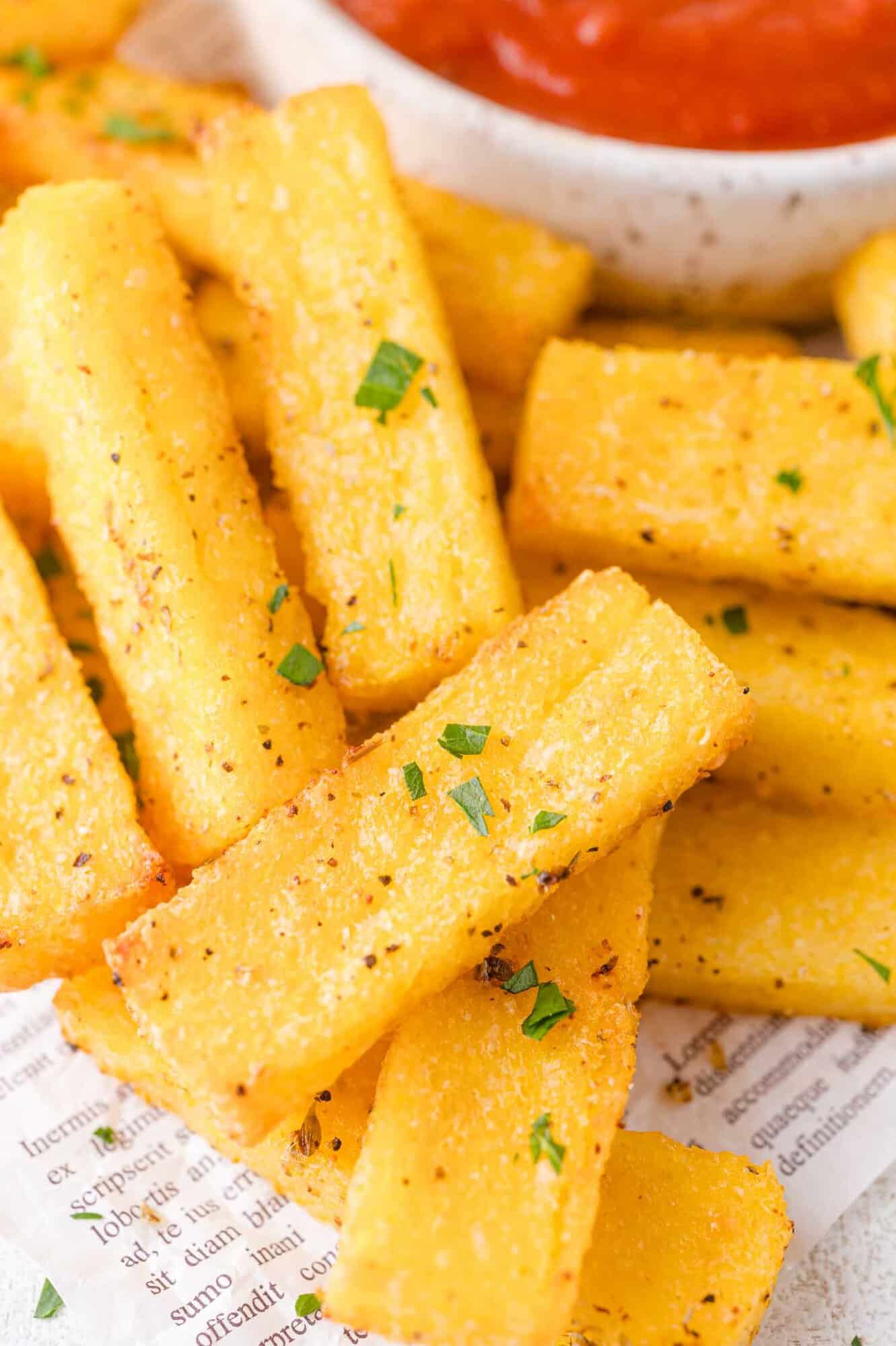 Golden polenta fries garnished with chopped herbs are arranged on paper, with a bowl of red dipping sauce in the background.
