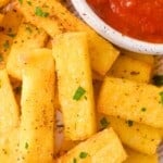 Rectangular pieces of seasoned polenta fries garnished with chopped herbs, served next to a bowl of red dipping sauce.