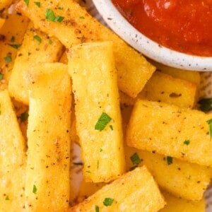 Rectangular pieces of seasoned polenta fries garnished with chopped herbs, served next to a bowl of red dipping sauce.