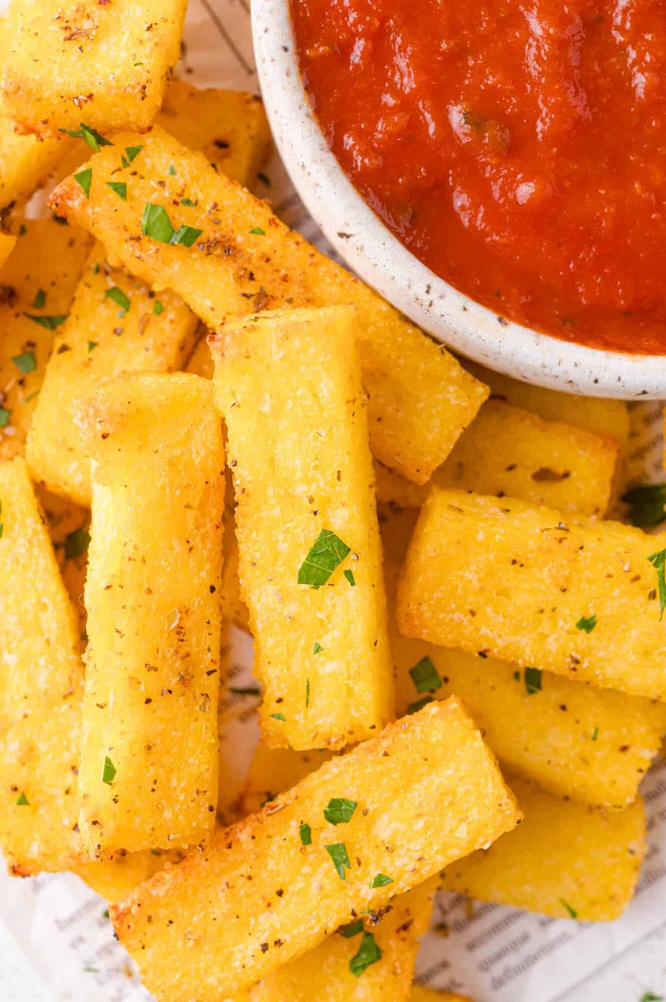 Golden polenta fries garnished with herbs are arranged next to a bowl of red marinara sauce.