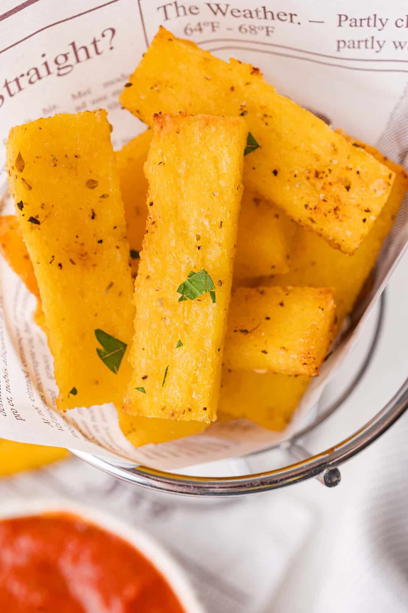 Crispy polenta fries seasoned with herbs, served in a metal basket lined with newspaper print paper, with a bowl of red dipping sauce nearby.