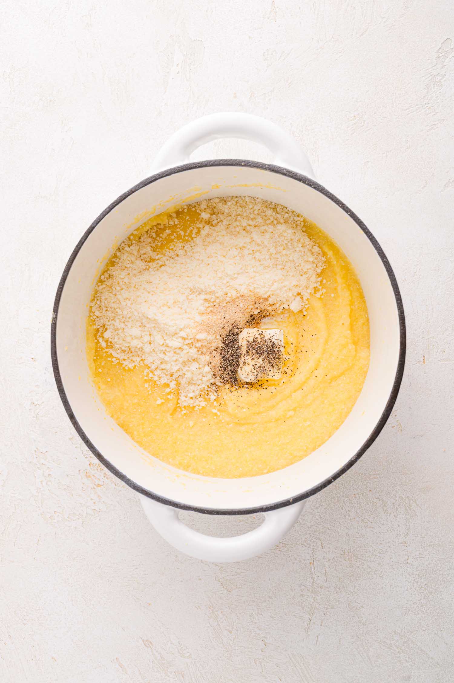 A white pot contains yellow polenta topped with shredded cheese, black pepper, and a pat of butter on a light-colored surface.