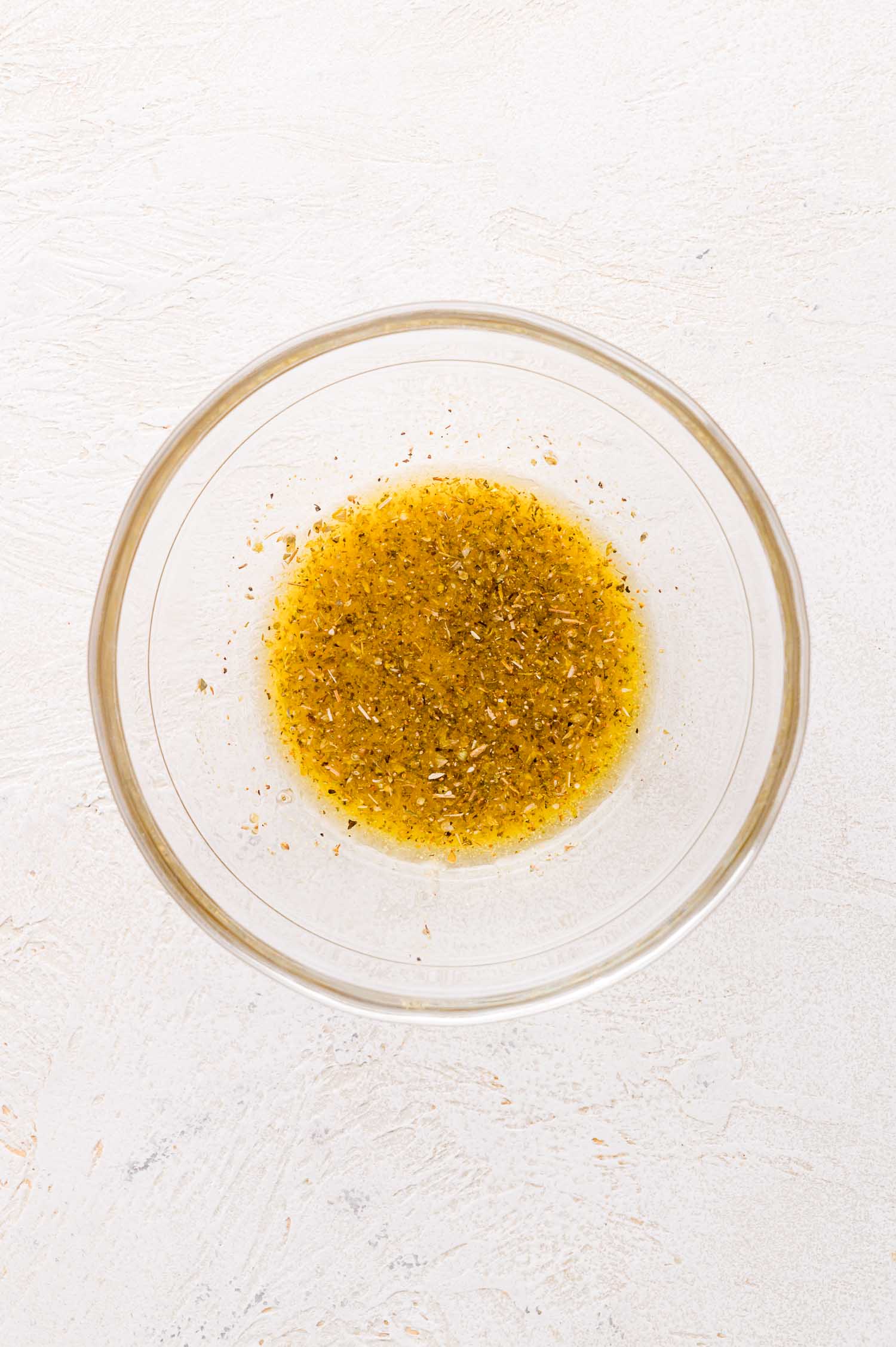 A clear glass bowl containing a yellowish olive oil mixed with dried herbs, placed on a light textured surface.