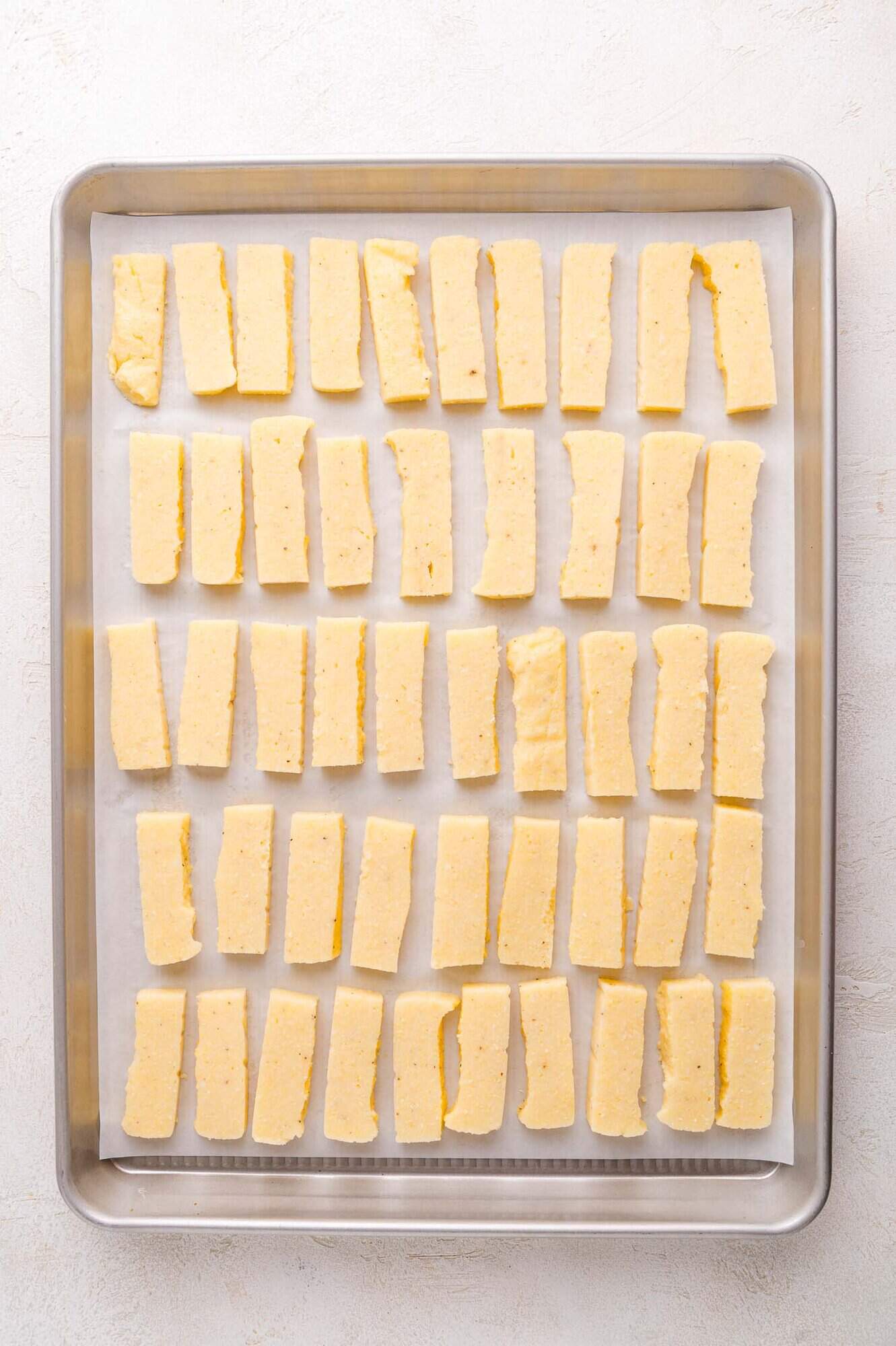 A baking sheet lined with parchment paper holds evenly spaced, rectangular strips of polenta arranged in rows.