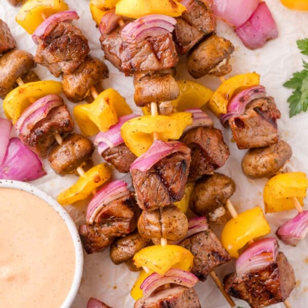 Grilled steak kabobs with yellow bell peppers, red onions, and mushrooms arranged on parchment paper beside a bowl of creamy dipping sauce.