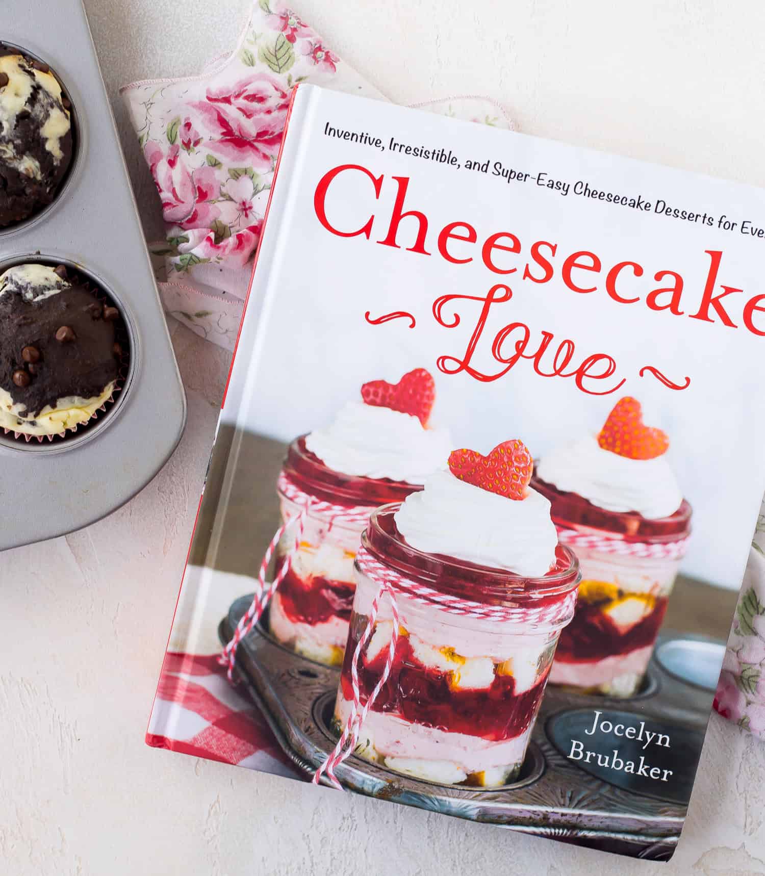 A cookbook titled Cheesecake Love by Jocelyn Brubaker is on a floral cloth next to a muffin tin with two baked muffins.