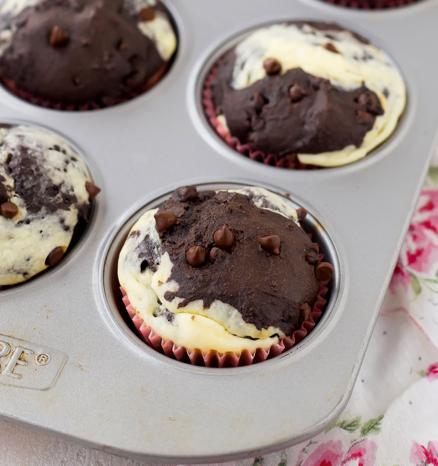 A muffin tin with marbled chocolate and cream cheese muffins, each topped with chocolate chips, on a floral-patterned cloth.