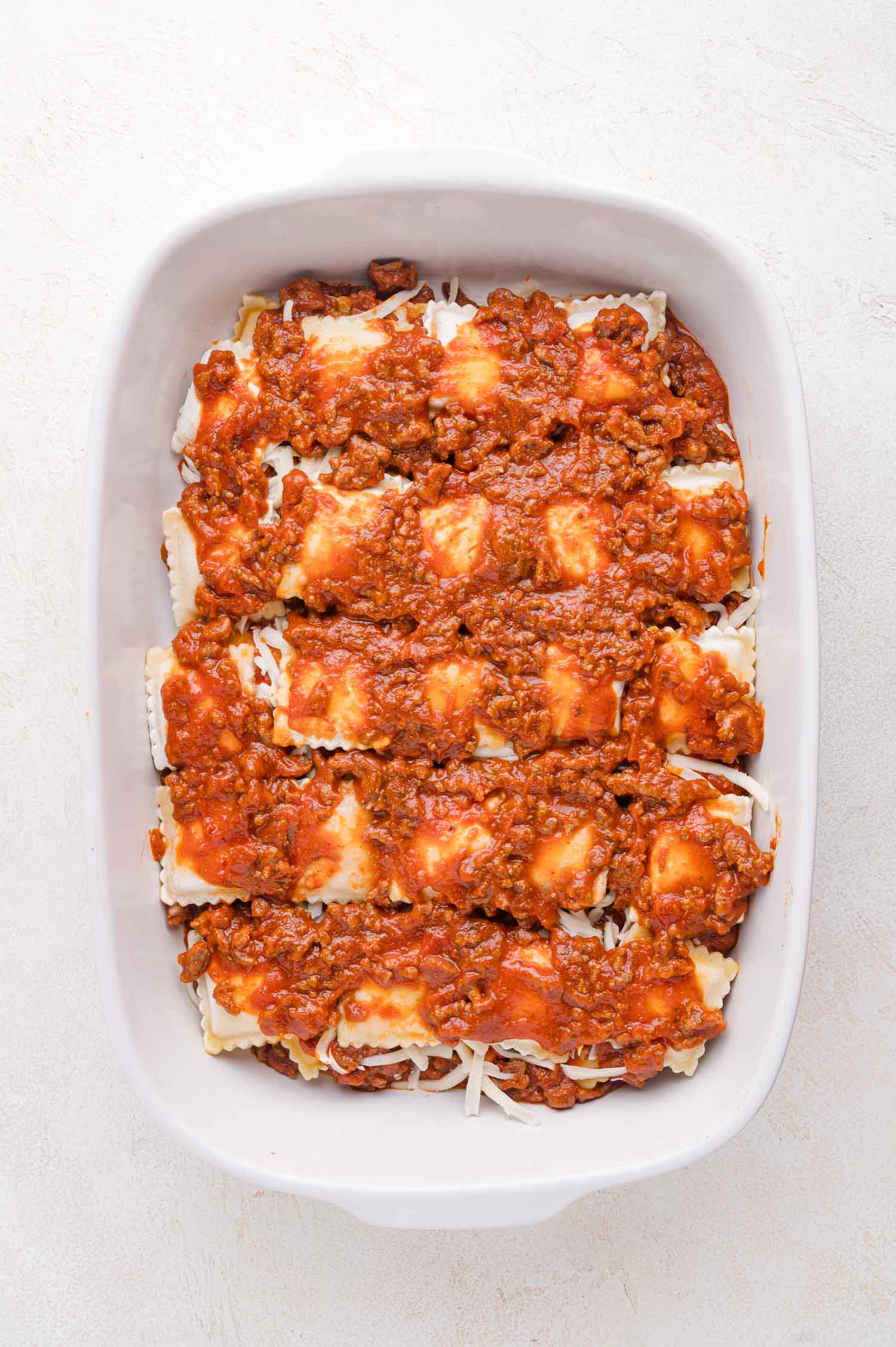 A white baking dish filled with layers of ravioli, shredded cheese, and meat sauce, ready to be baked.