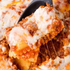 A close-up of a serving spoon lifting a cheesy, meat-filled ravioli from a baked pasta dish topped with melted cheese and tomato sauce.