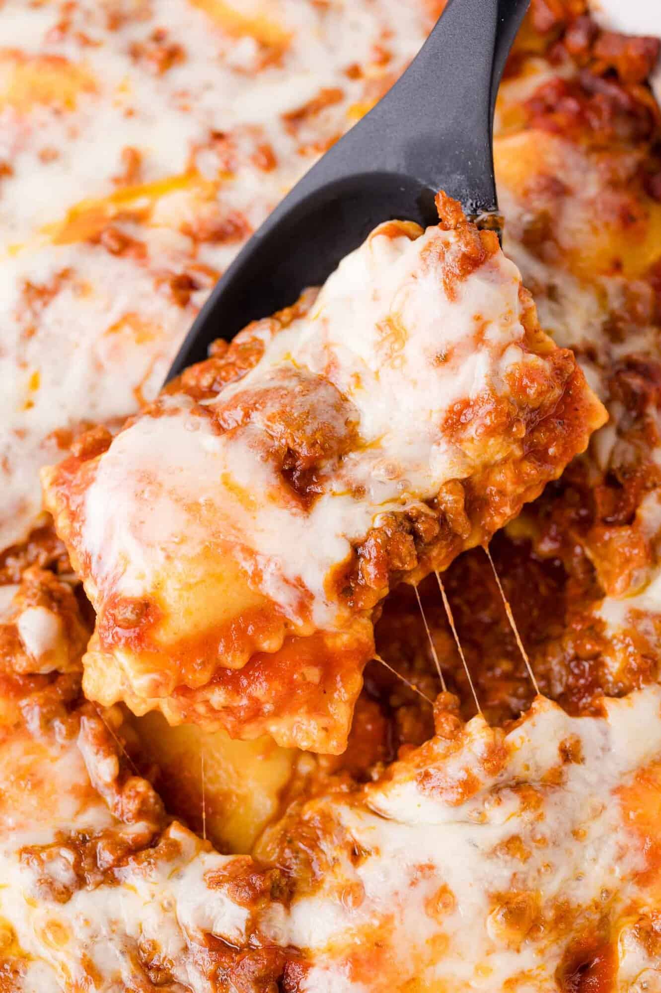 A close-up of a serving spoon lifting a cheesy, meat-filled ravioli from a baked pasta dish topped with melted cheese and tomato sauce.