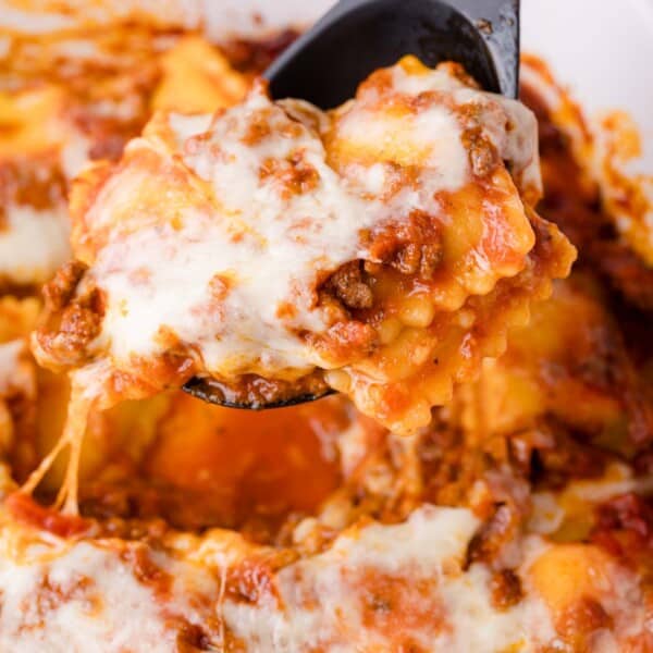 A close-up of a spatula lifting a cheesy, baked ravioli dish with tomato sauce and ground meat.