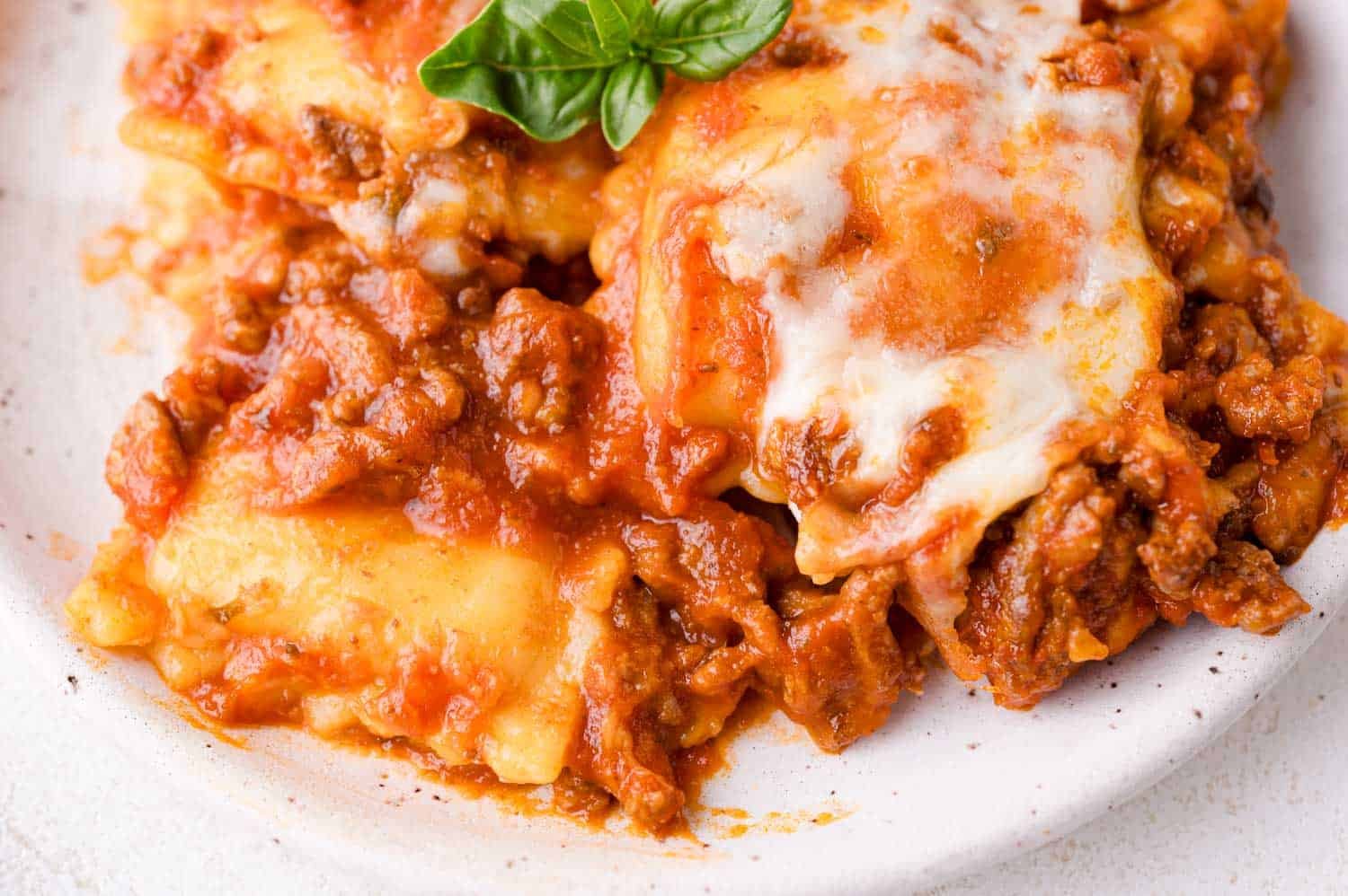 Close-up of a serving of baked ravioli topped with melted cheese and tomato meat sauce, garnished with a fresh basil leaf on a white plate.