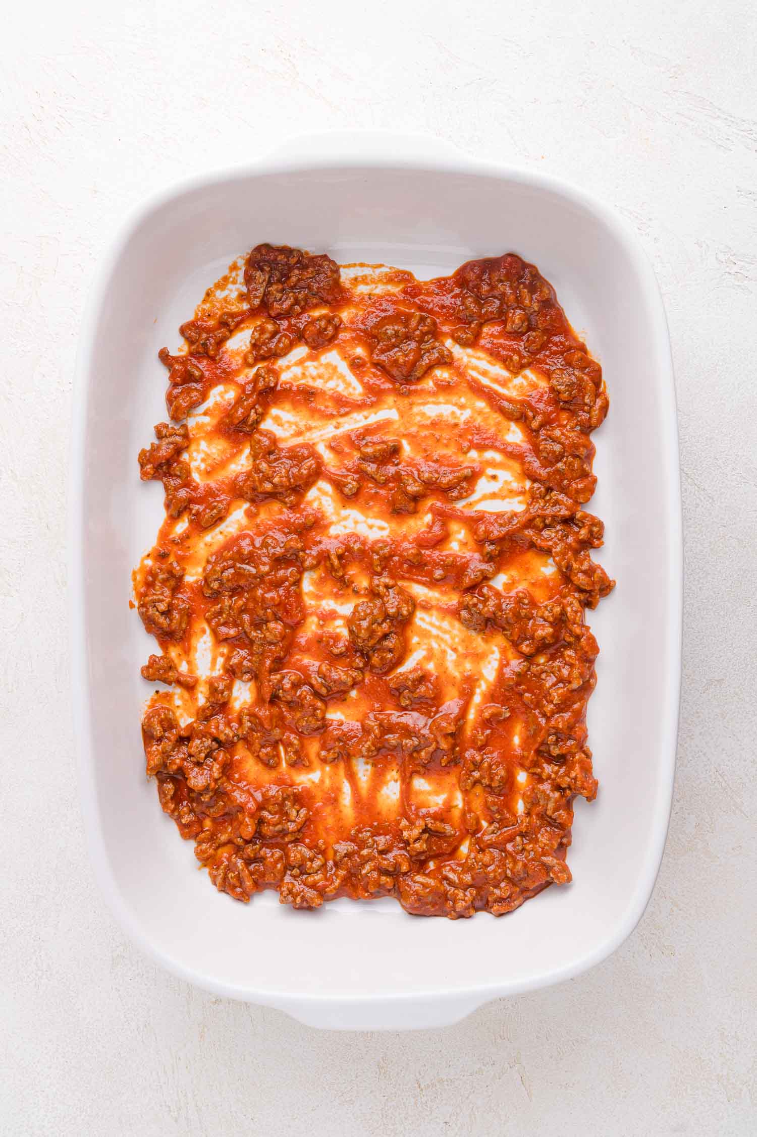 A white casserole dish with a layer of cooked ground beef in tomato sauce spread across the bottom.