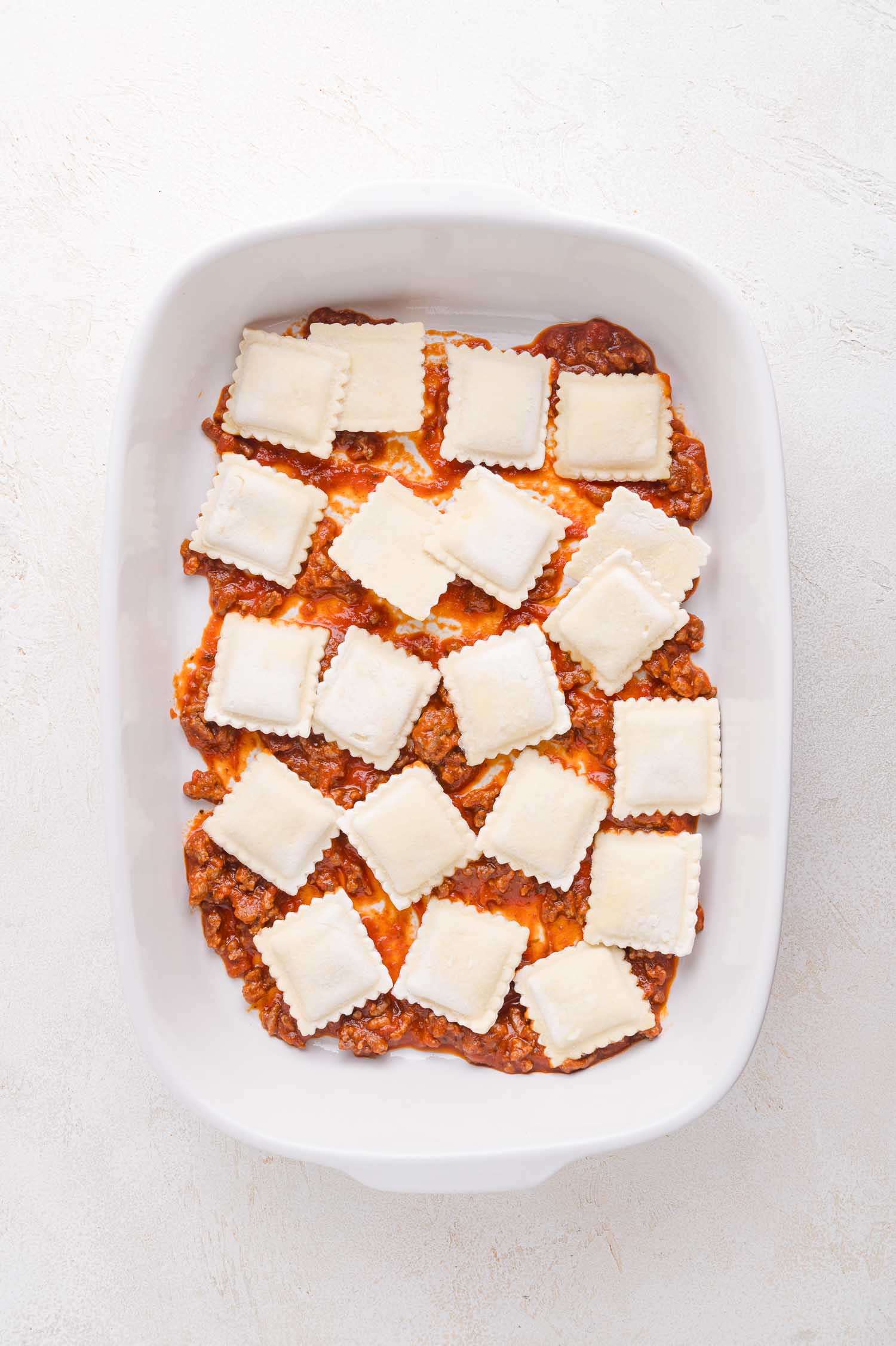 A white baking dish with a layer of meat sauce and uncooked ravioli arranged evenly on top.