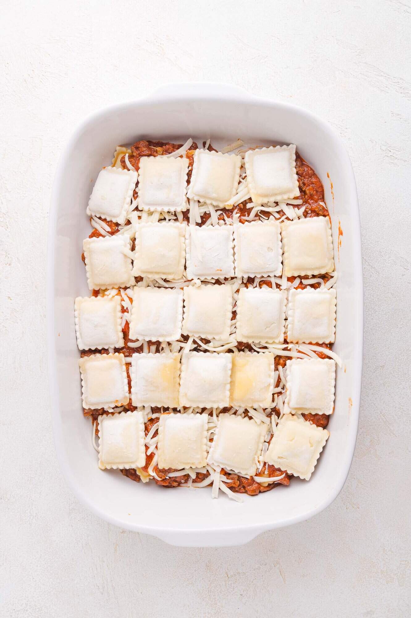 A white baking dish filled with a layer of ground meat sauce, shredded cheese, and uncooked square ravioli arranged in rows.
