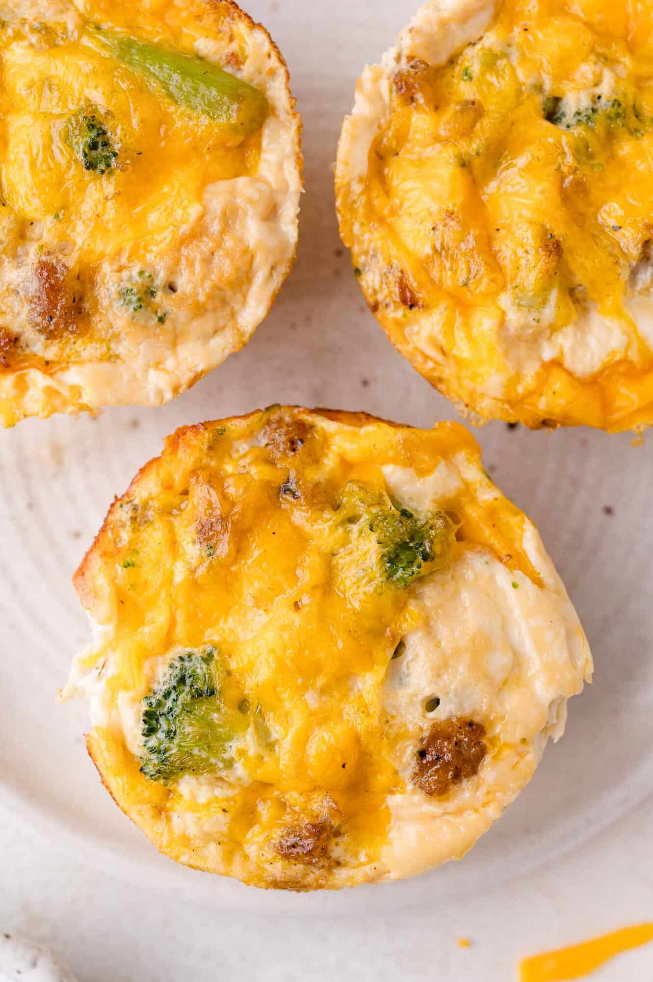 Three baked egg white muffins with visible broccoli, sausage, and melted cheddar cheese on a white plate.