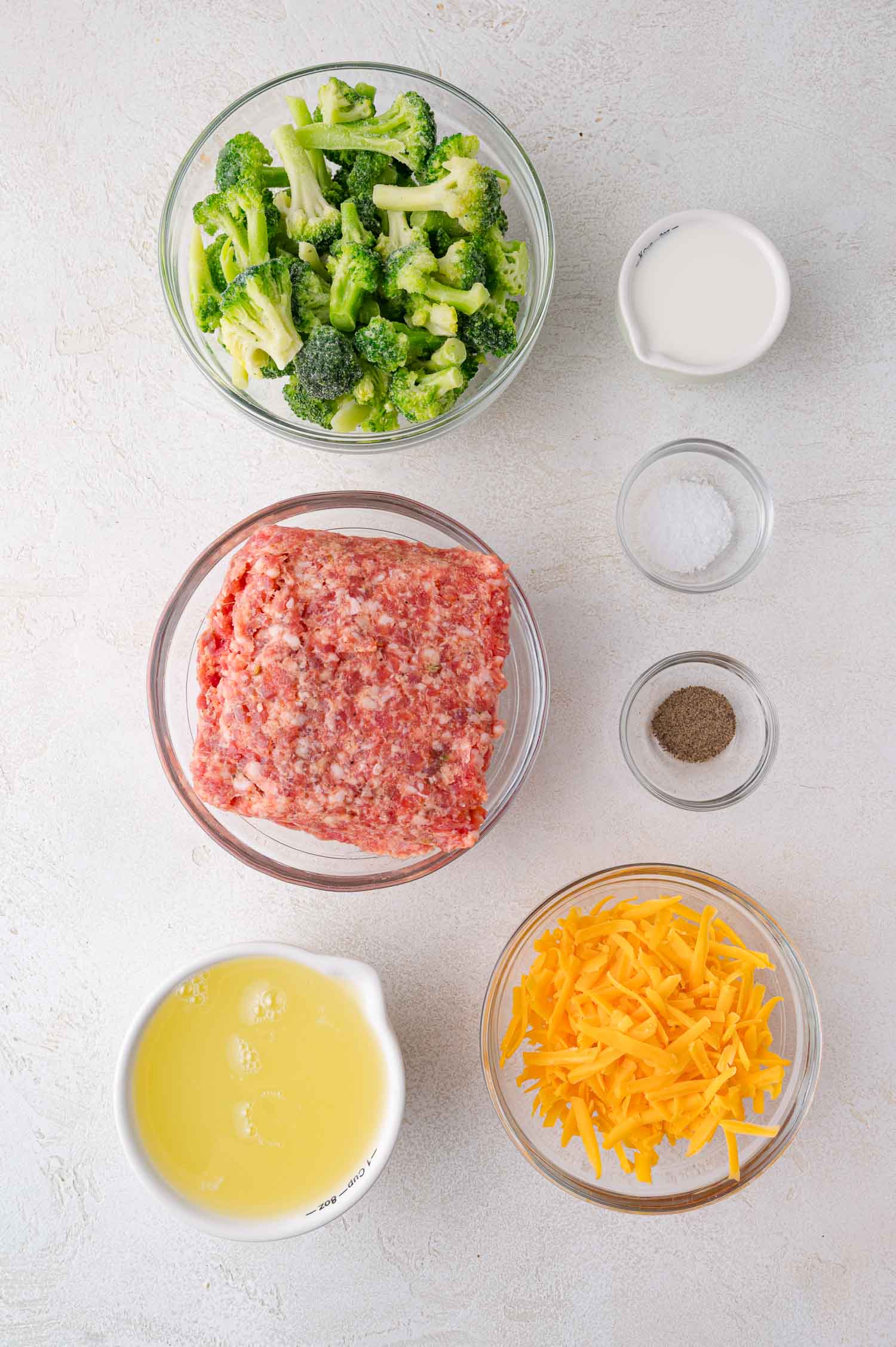Bowls containing chopped broccoli, ground sausage, shredded cheddar cheese, egg whites, milk, salt, and pepper are arranged on a white surface.