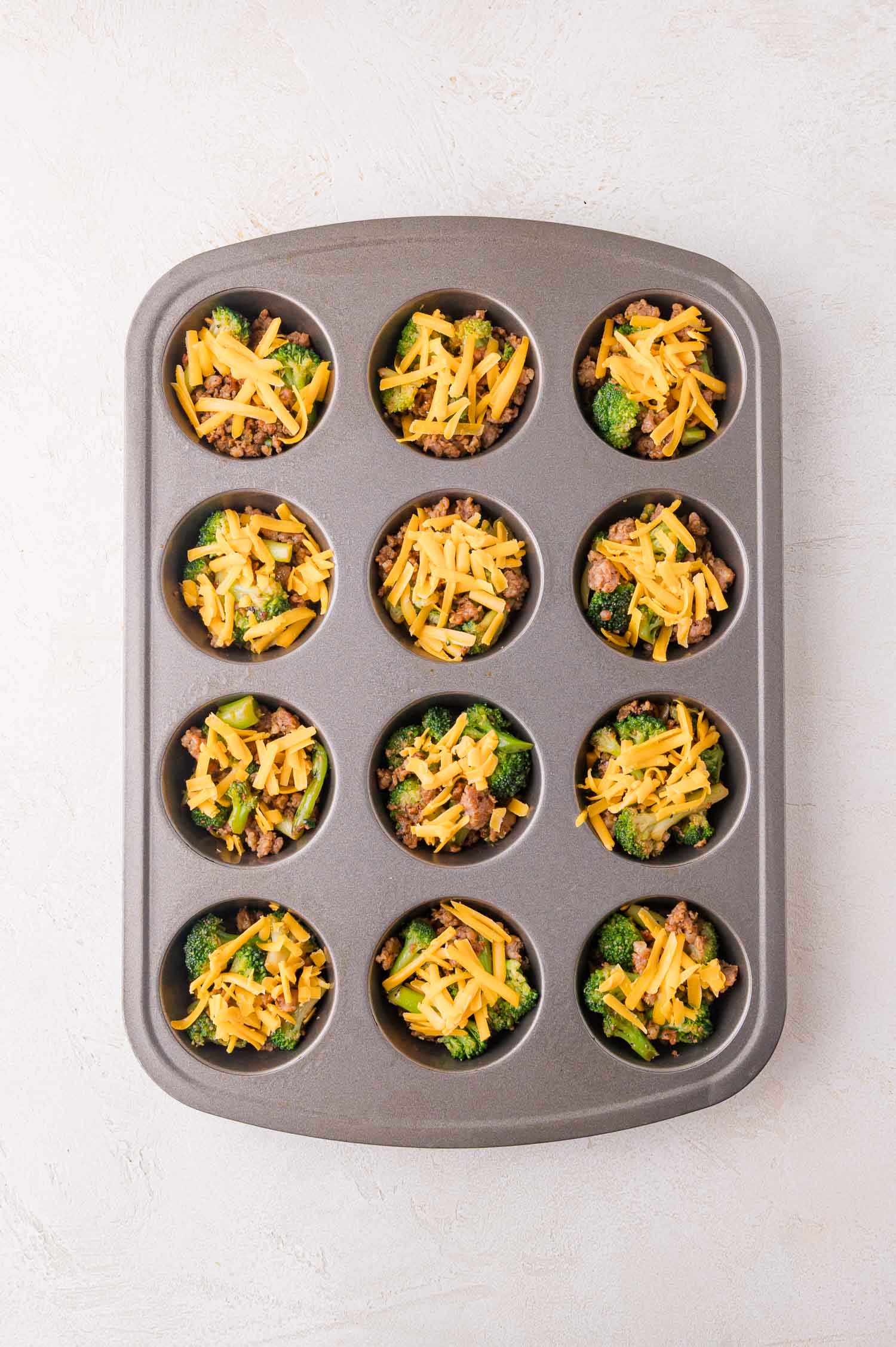 A muffin tin with twelve cups, each filled with broccoli, ground sausage, and shredded cheddar cheese, on a light-colored surface.