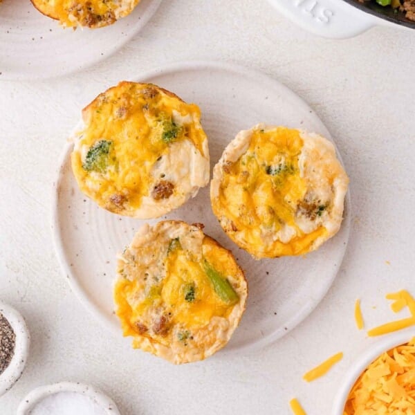 Three egg white muffins with melted cheese and visible broccoli pieces are arranged on a white plate, surrounded by ingredients and kitchen items on a light countertop.