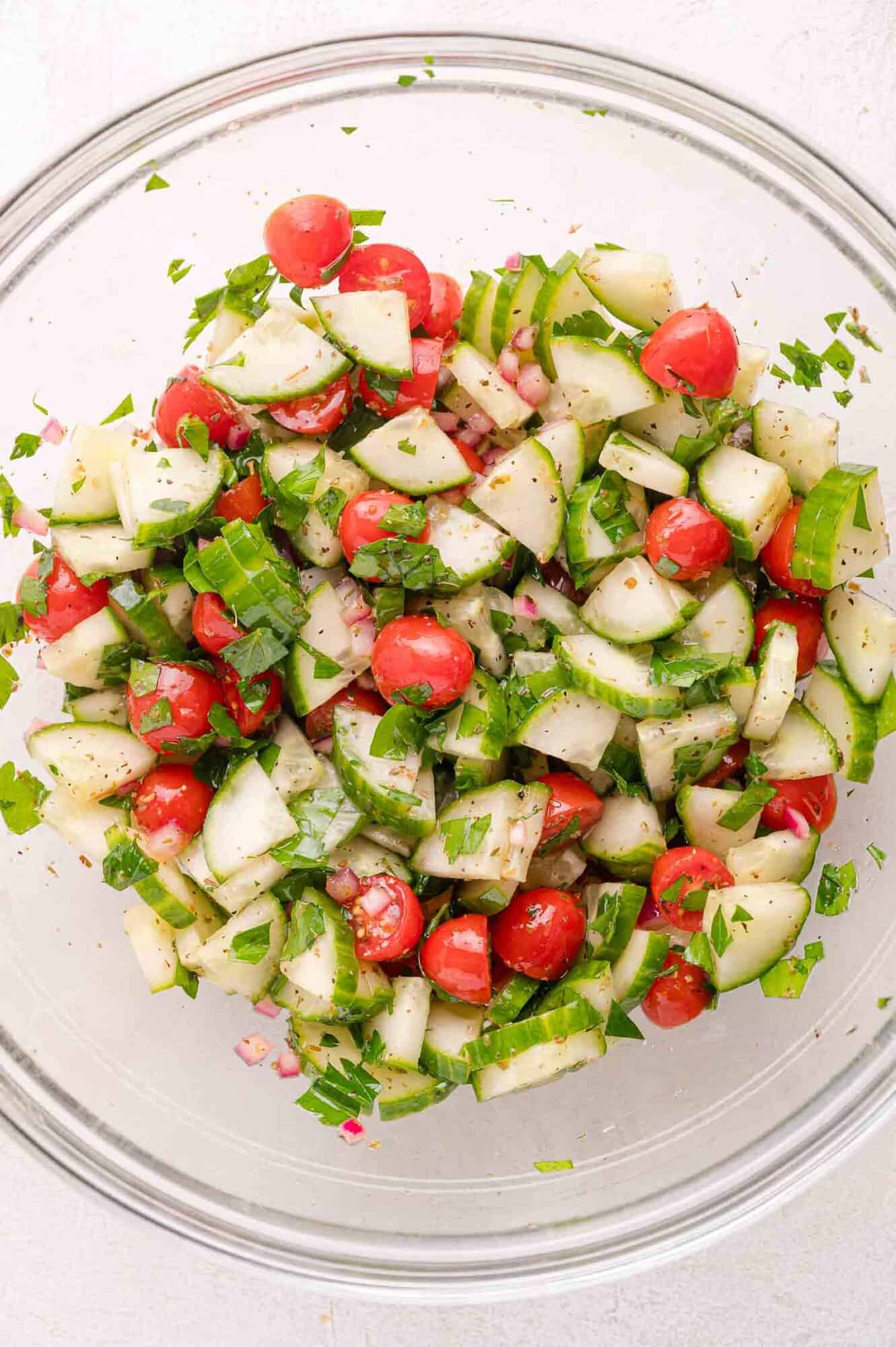 A glass bowl with chopped cucumbers, cherry tomatoes, red onions, and parsley.