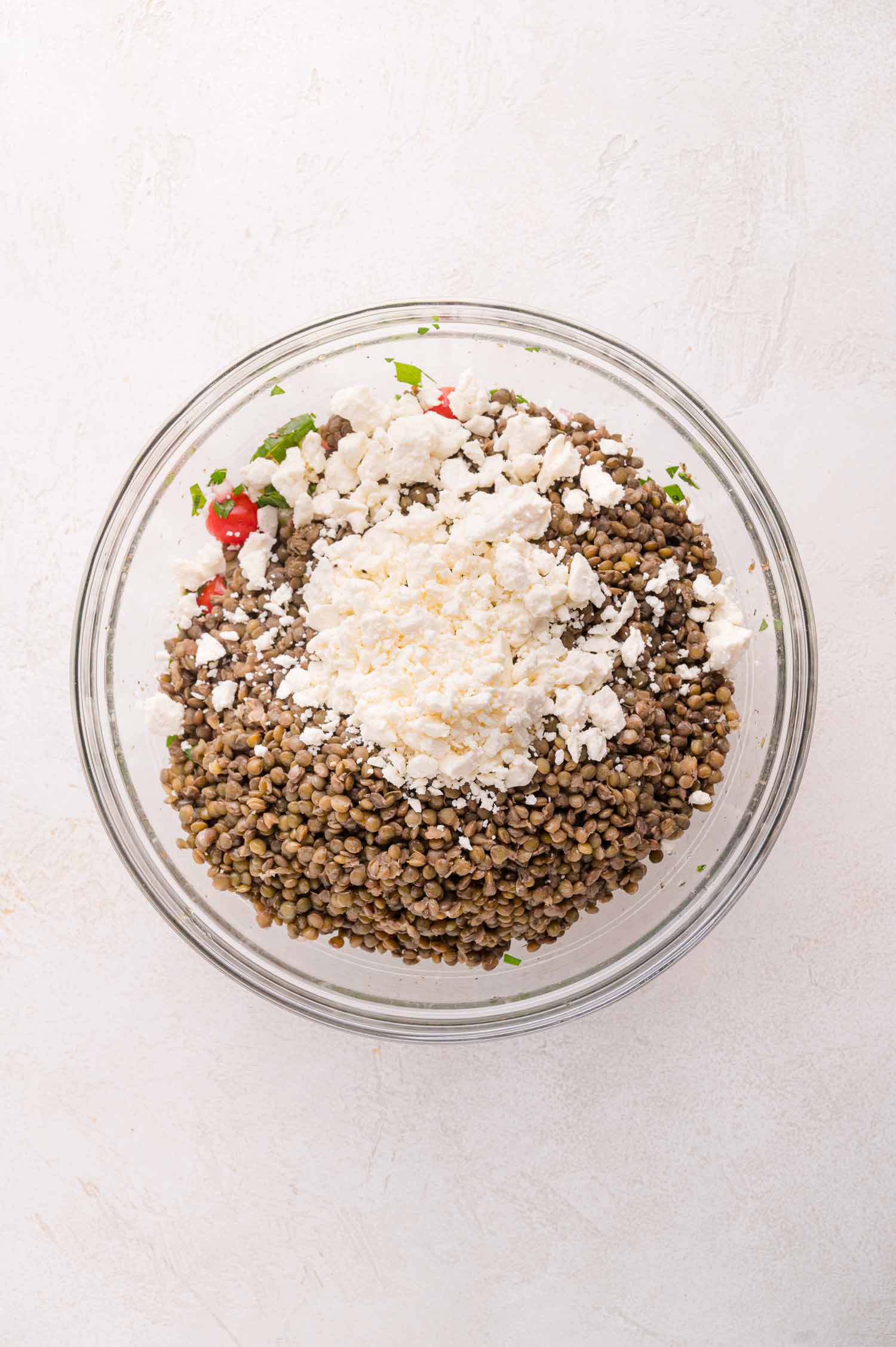 A glass bowl filled with lentils, chopped vegetables, and a generous amount of crumbled white cheese on top, placed on a light-colored surface.