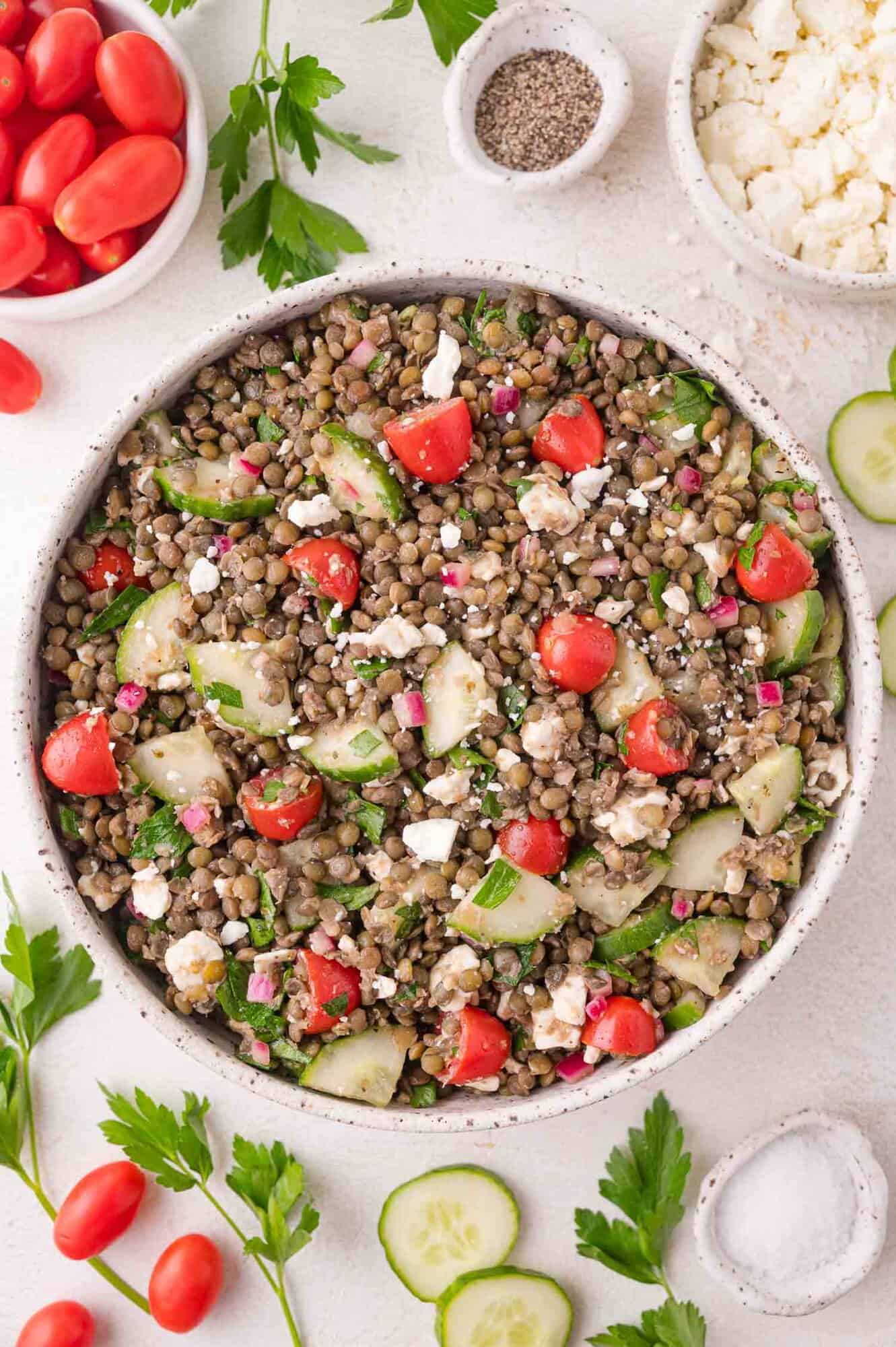 A bowl of lentil salad with cherry tomatoes, cucumber slices, red onion, parsley, and crumbled feta cheese, surrounded by fresh ingredients on a white surface.