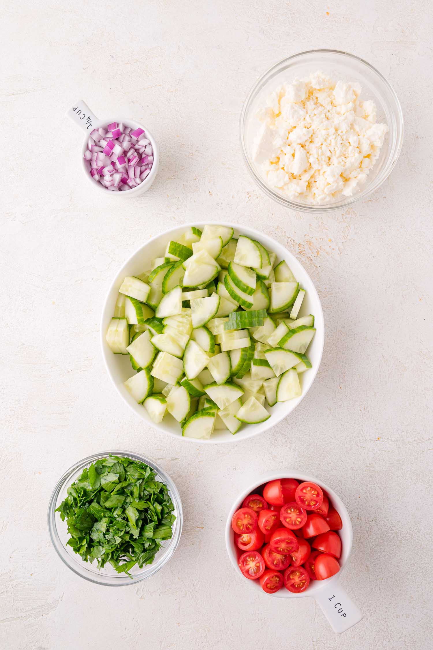 Five bowls containing chopped red onion, crumbled feta cheese, sliced cucumber, halved cherry tomatoes, and chopped parsley are arranged on a light surface.