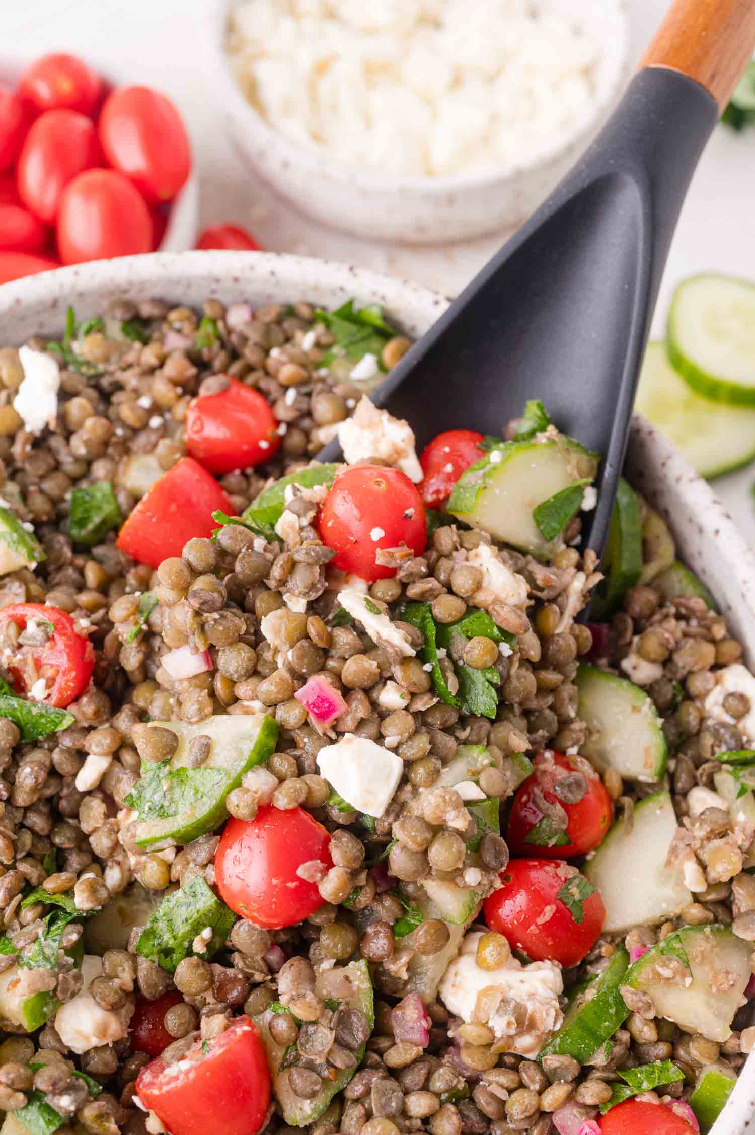 A bowl of lentil salad with cherry tomatoes, cucumber, feta cheese, red onion, and parsley, with a black serving spoon.