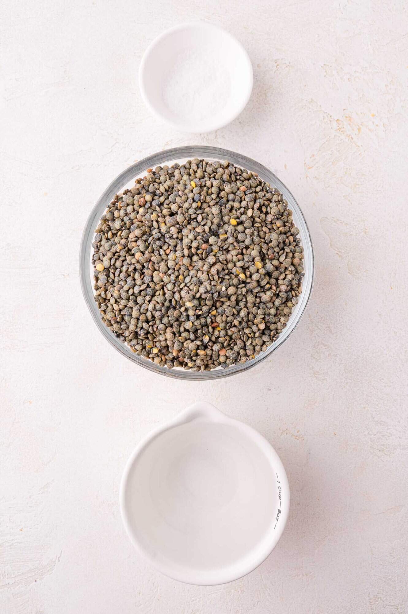 A bowl of dry lentils, a small bowl of salt, and an empty measuring cup arranged on a light-colored surface.