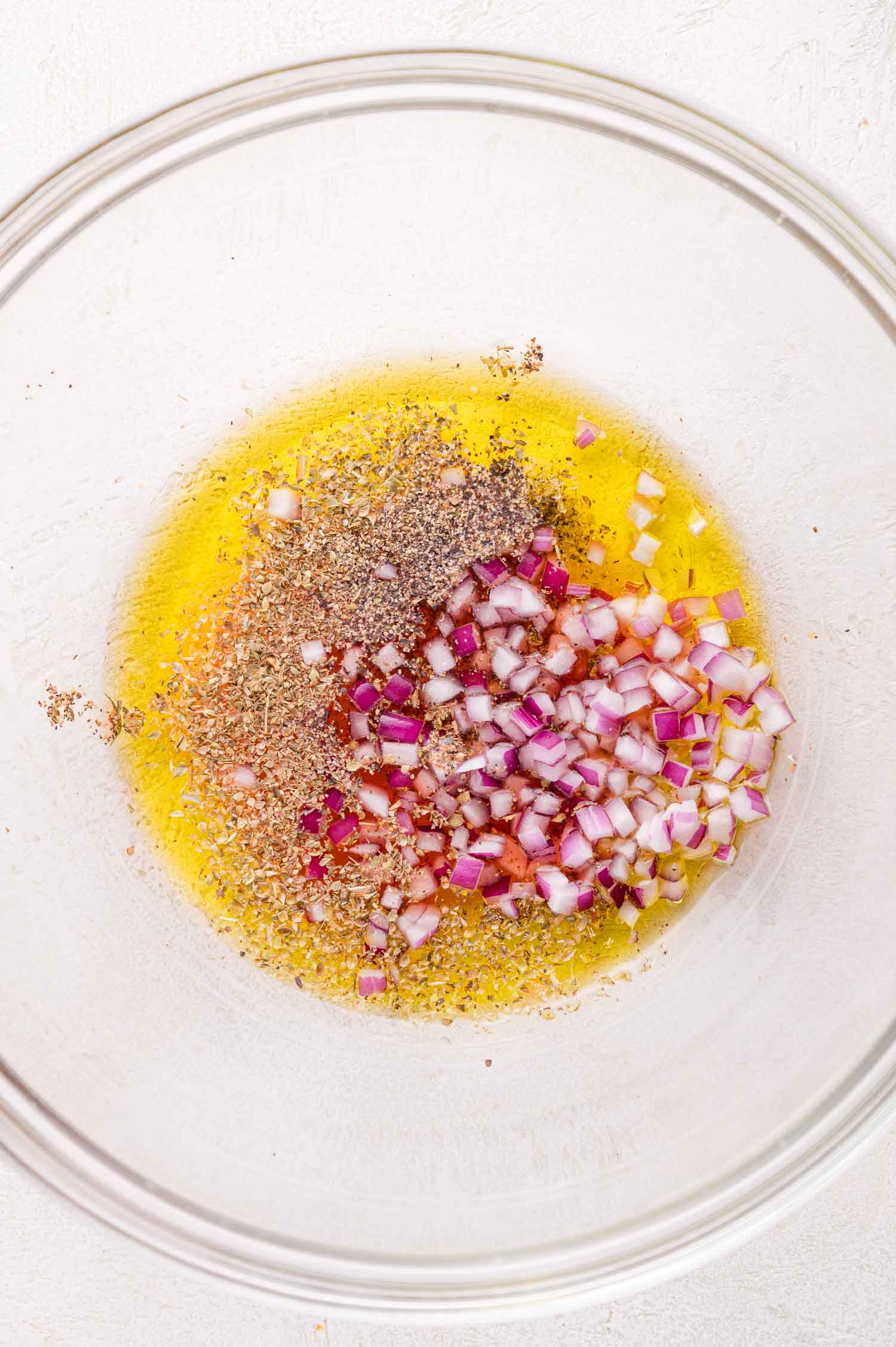 A glass bowl containing olive oil, dried herbs, and chopped red onion on a light-colored surface.