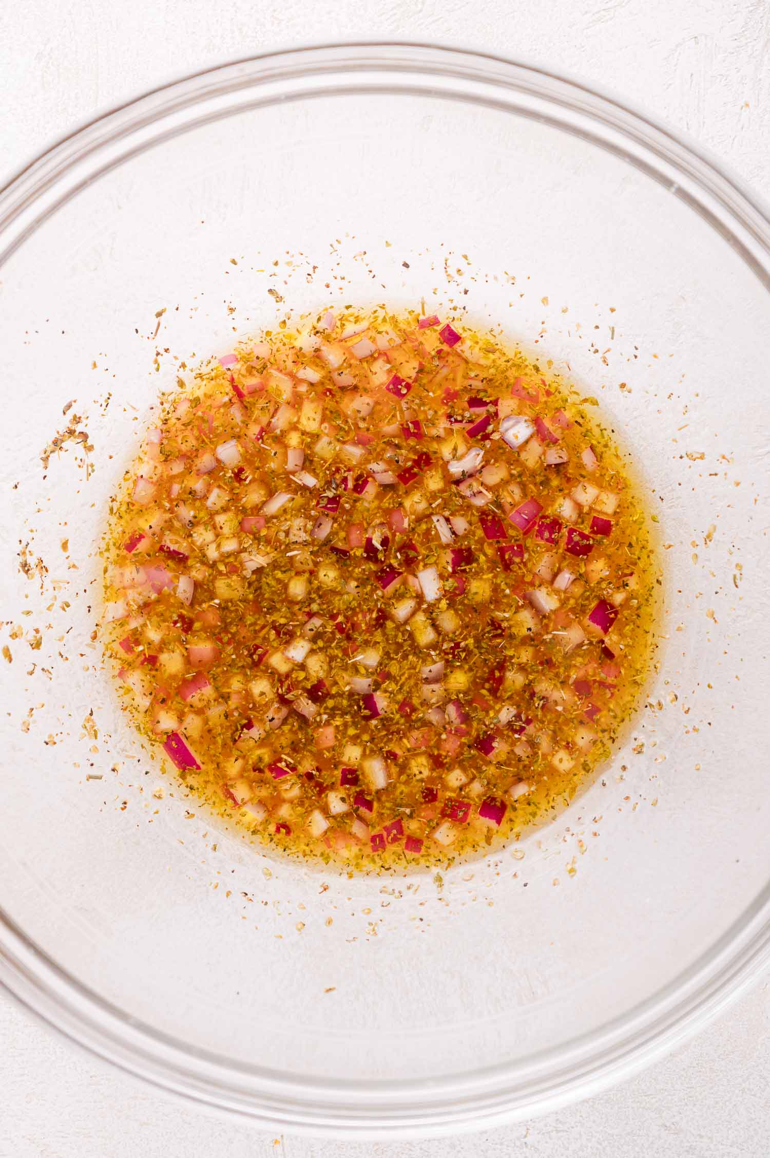 A clear glass bowl containing a mixture of olive oil, red onion, and dried herbs on a white surface.