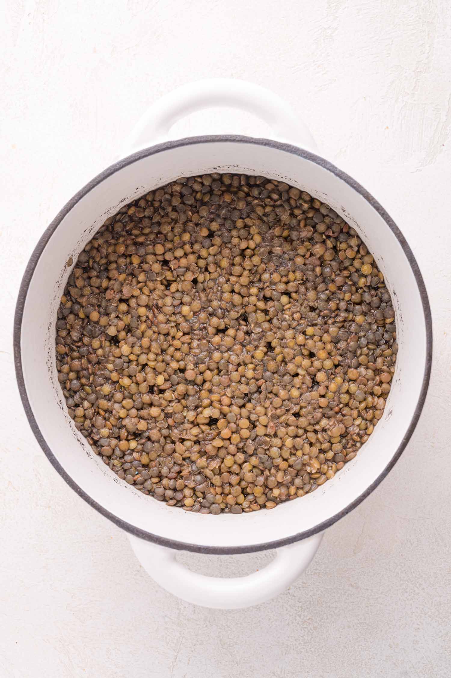 A white pot filled with cooked brown lentils viewed from above on a light-colored surface.