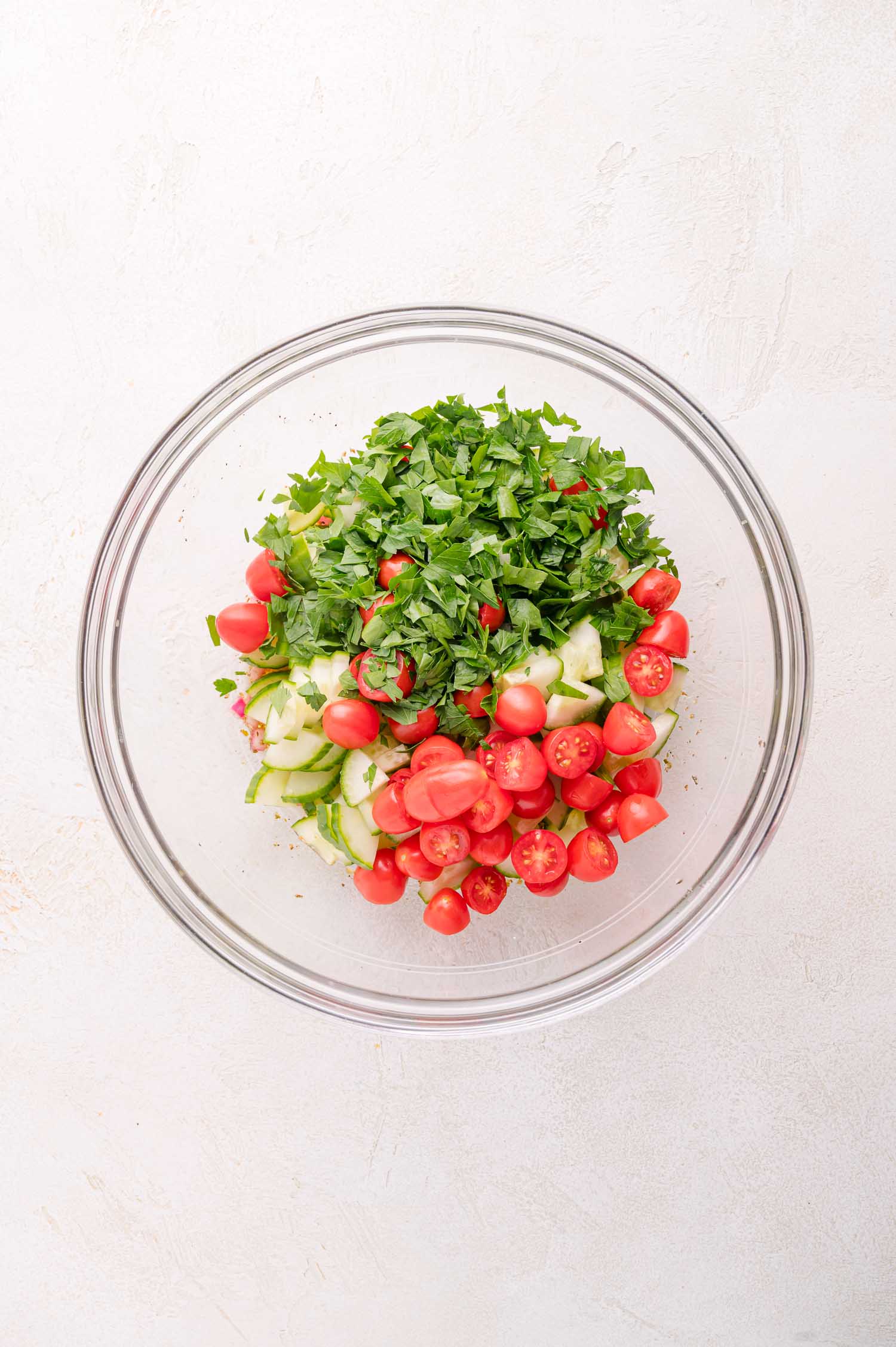 A glass bowl containing chopped tomatoes, cucumbers, and parsley on a white surface.