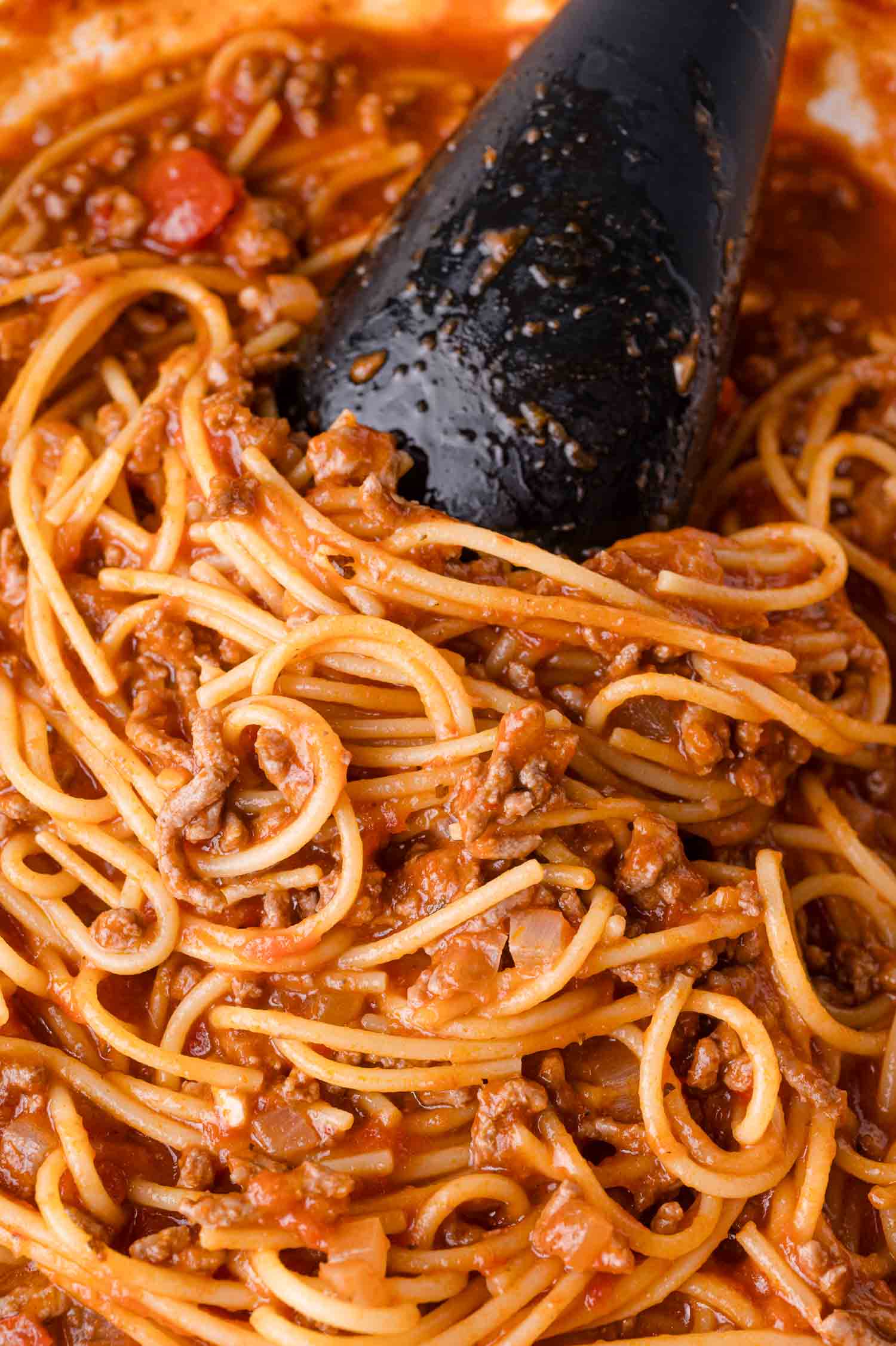 Close-up of cooked spaghetti mixed with ground meat and tomato sauce, with a black serving spoon partially visible.