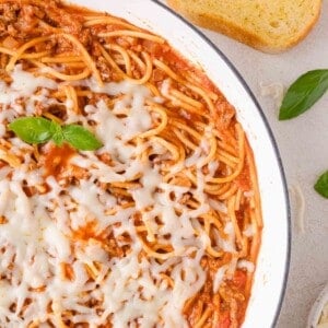 A skillet of one pan spaghetti topped with melted cheese, garnished with basil, sits on a light surface with a slice of bread nearby.