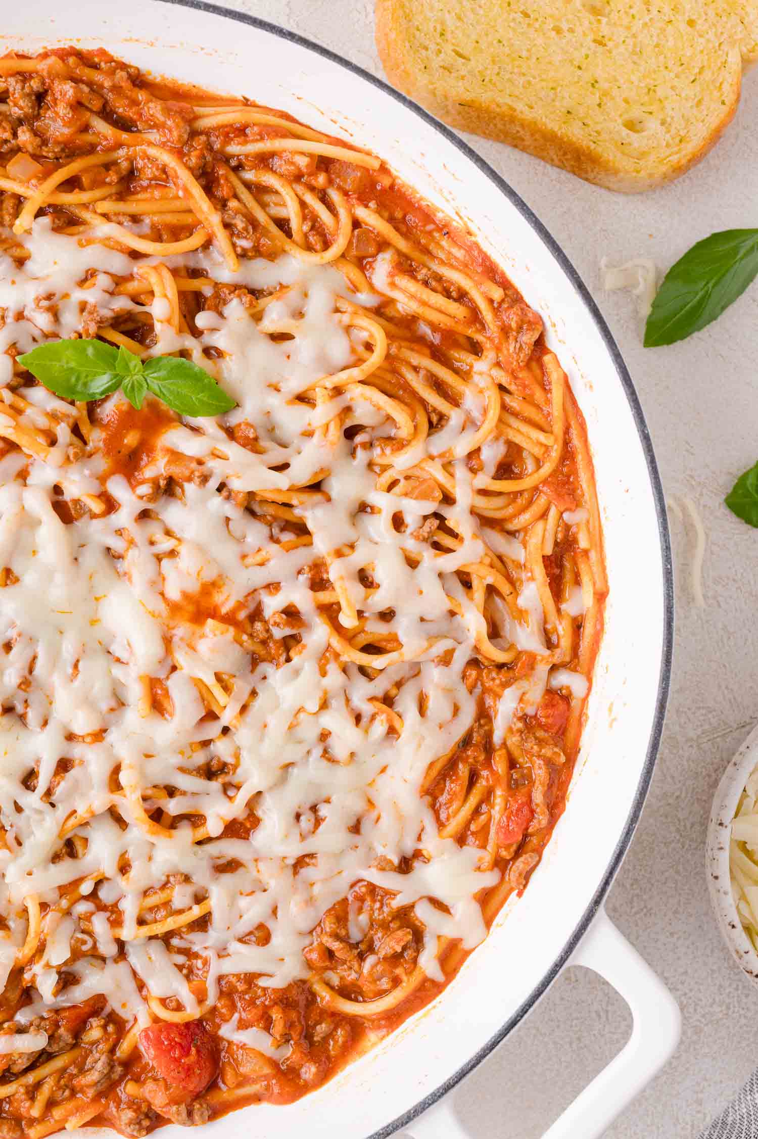 Spaghetti with meat sauce topped with melted cheese in a white pan, garnished with basil, with a slice of garlic bread and basil leaves on the side.