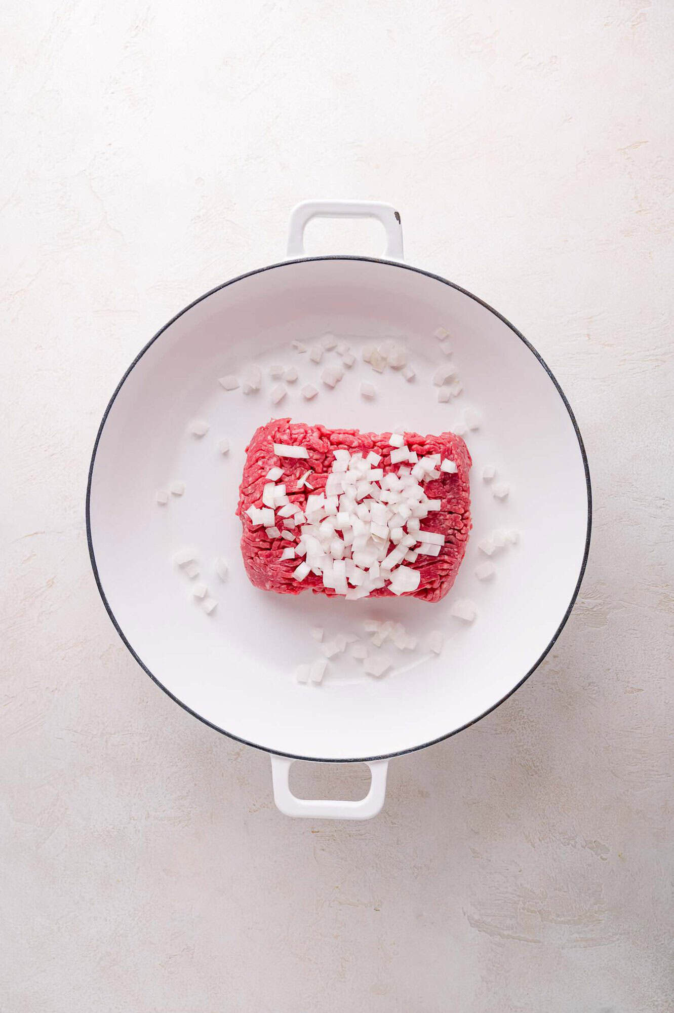 A block of raw ground beef and chopped onions in a white pot on a light-colored surface.