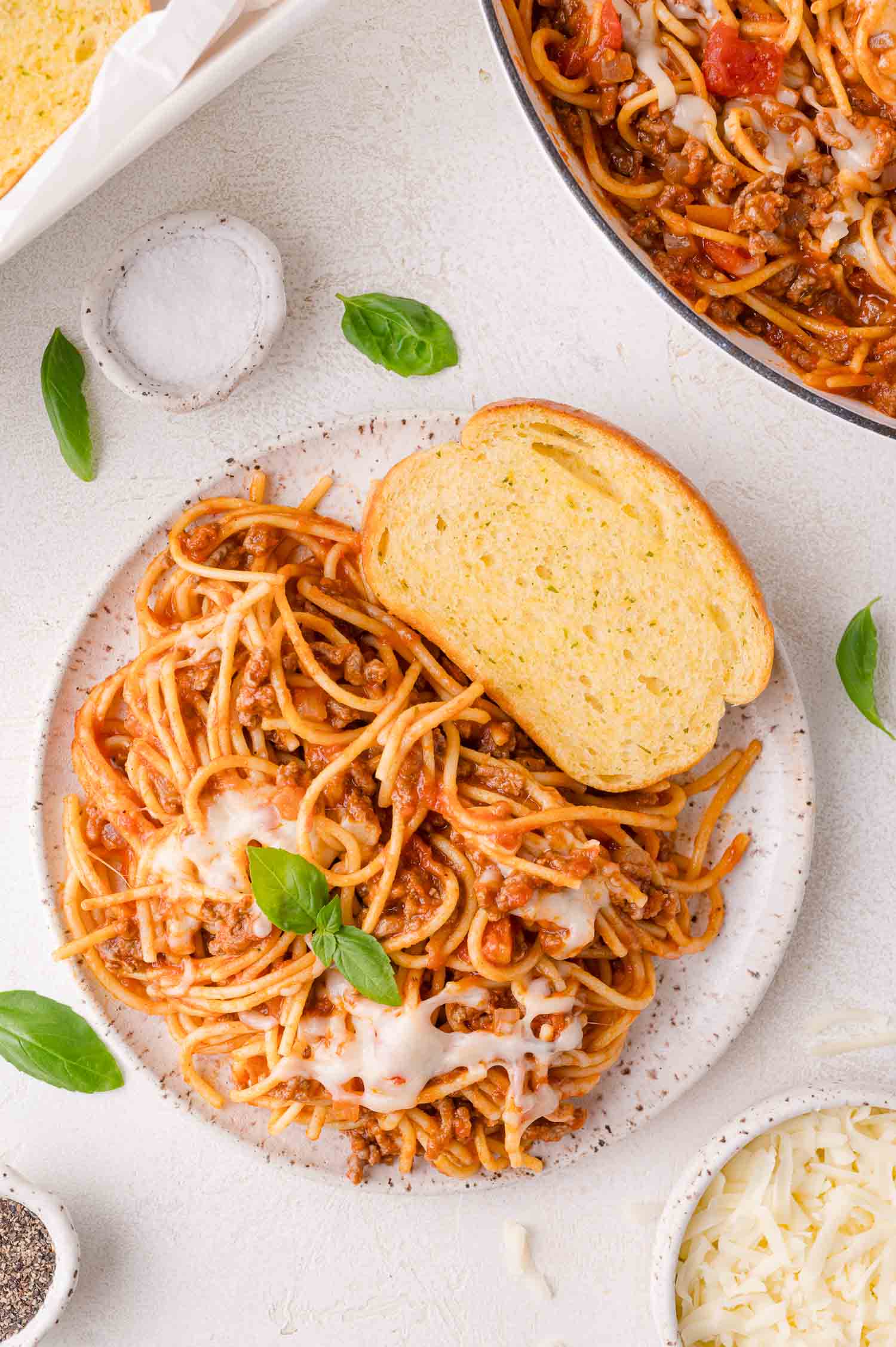 A plate of spaghetti with meat sauce and melted cheese, garnished with basil, served with two slices of garlic bread on a white surface.